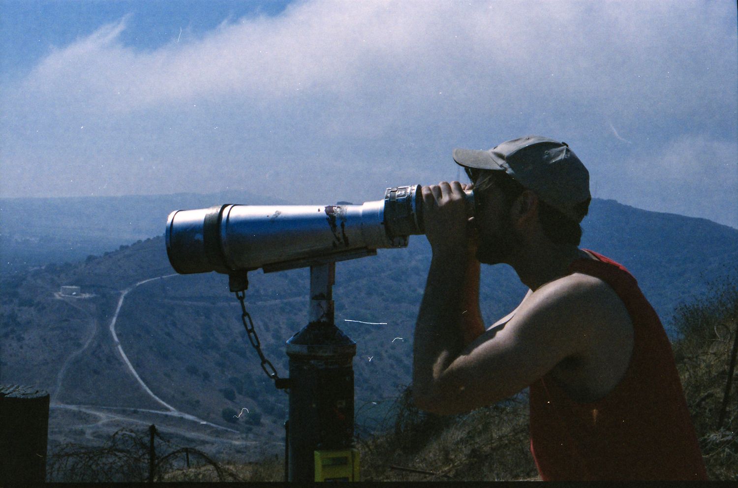Man using large telescope binoculars on mountain overlook with scenic winding road and hills in background.