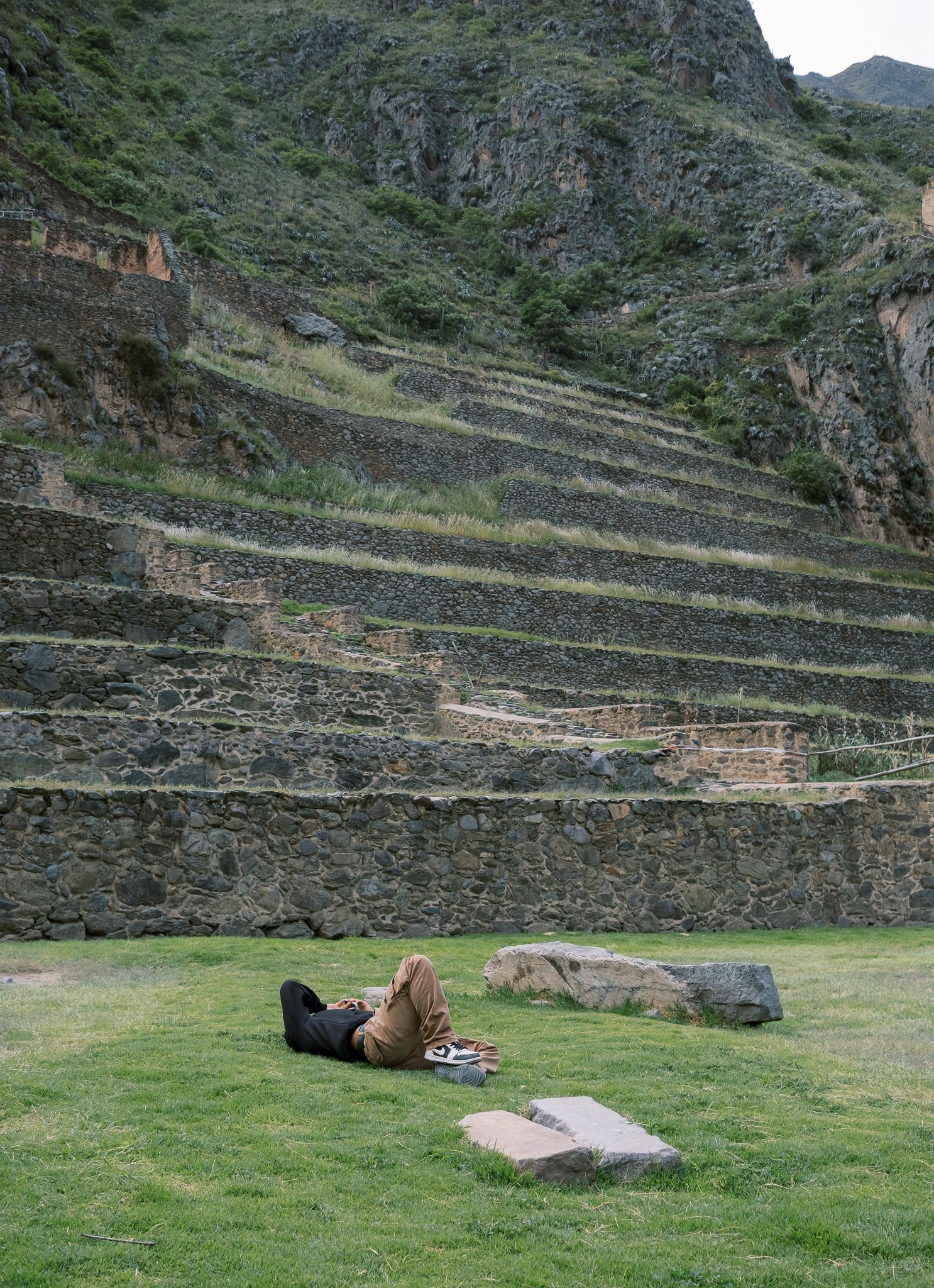Person relaxing on green grass beside ancient Incan stone terraces on a lush mountainside at Ollantaytambo, Peru.