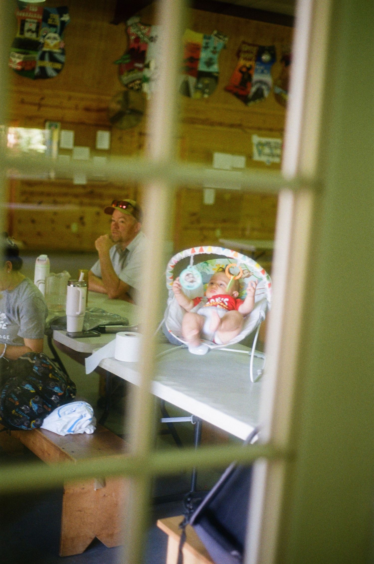 Baby resting in a colorful bouncer seat on a table inside a rustic room with an adult sitting nearby.