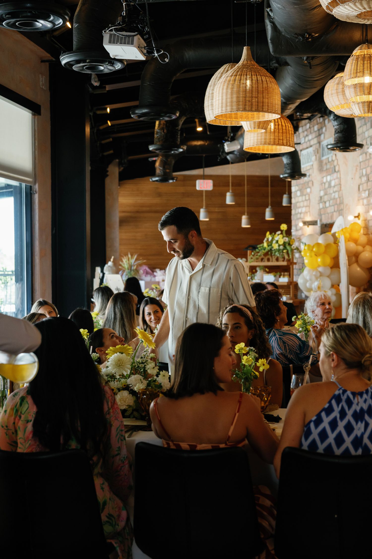 An indoor gathering space with rattan pendant lights and exposed brick walls filled with party guests.