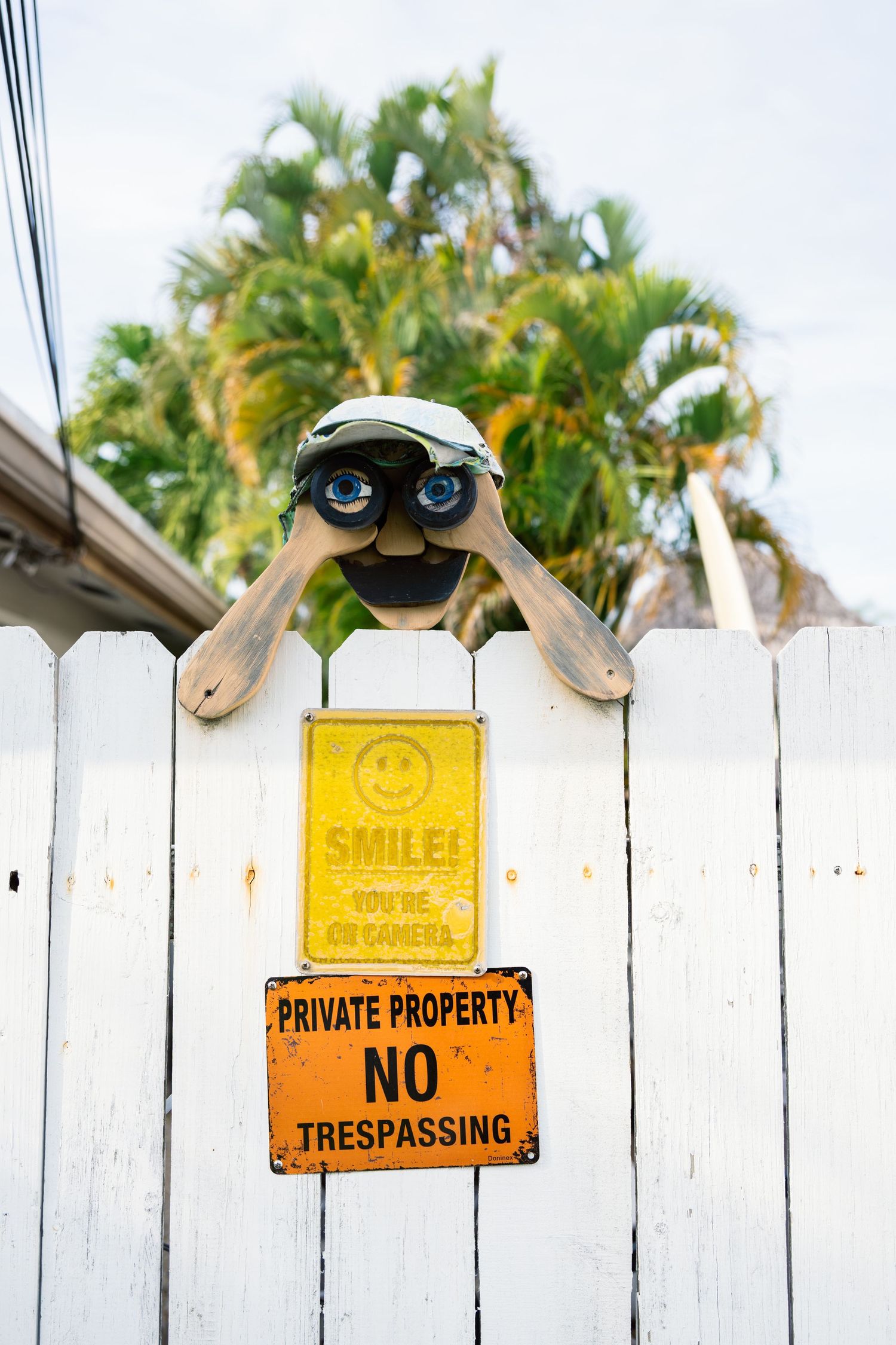 White picket fence with yellow smile sign and no trespassing warning against palm tree backdrop.