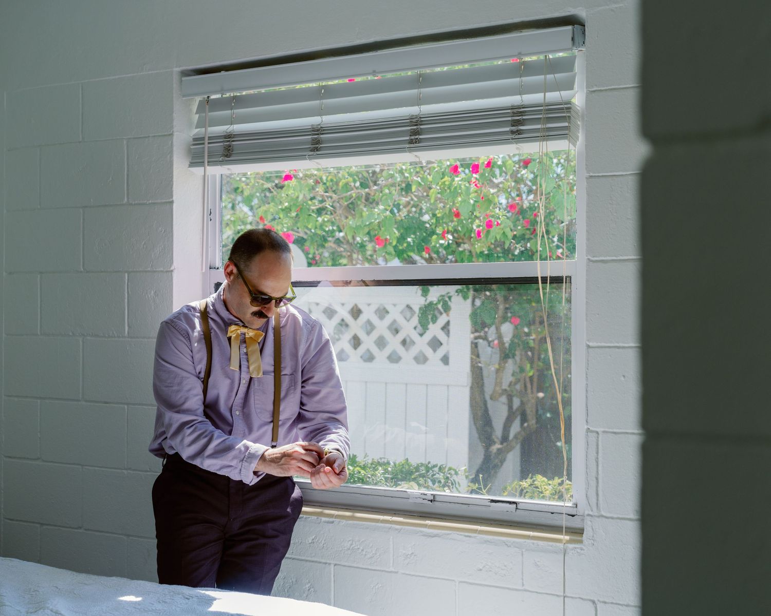 A person in a purple jacket sits at a window with green tiled walls and floral curtains.