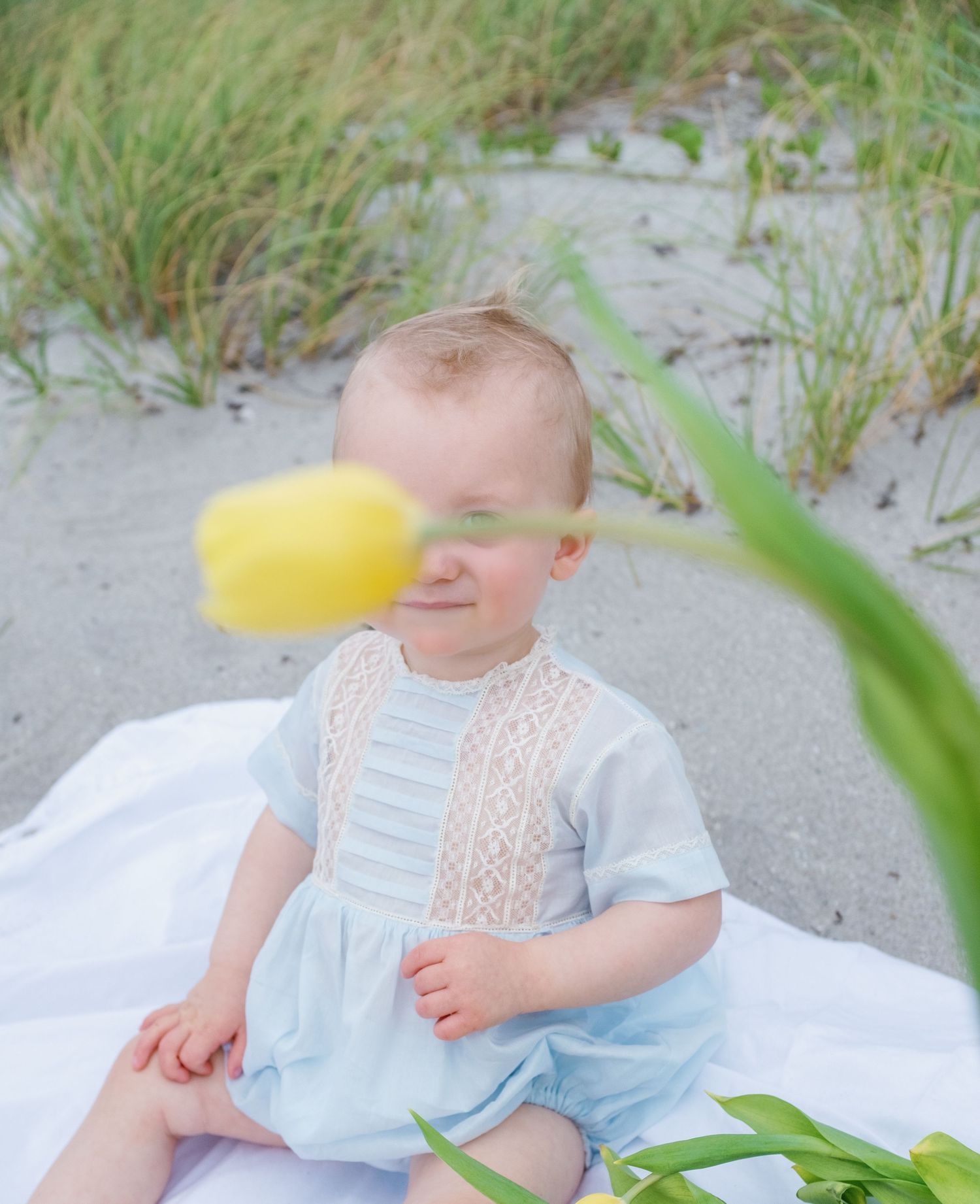 Child in blue and white dress sitting on sandy beach with sea grass in foreground.