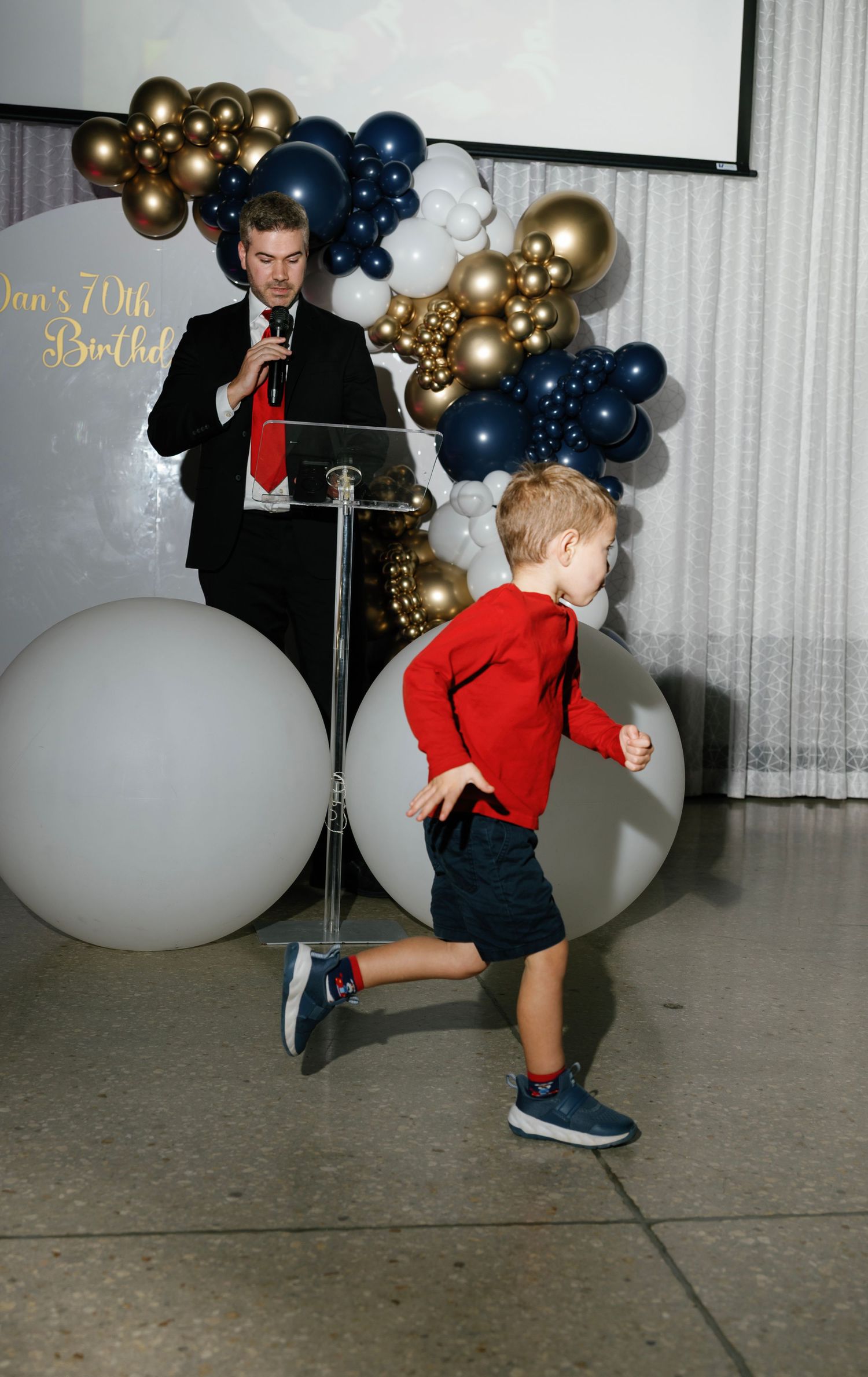 Young child in red dancing and moving across the floor at a party with gold and navy balloon decor.