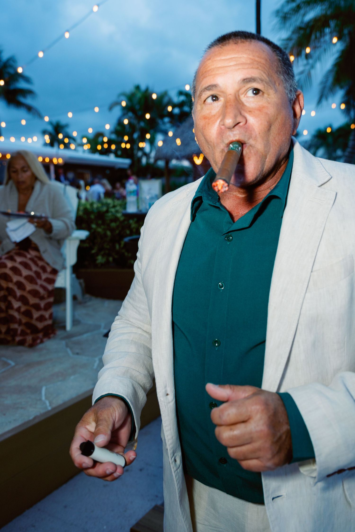 Man in light blazer and dark shirt enjoying cigars at outdoor evening reception.