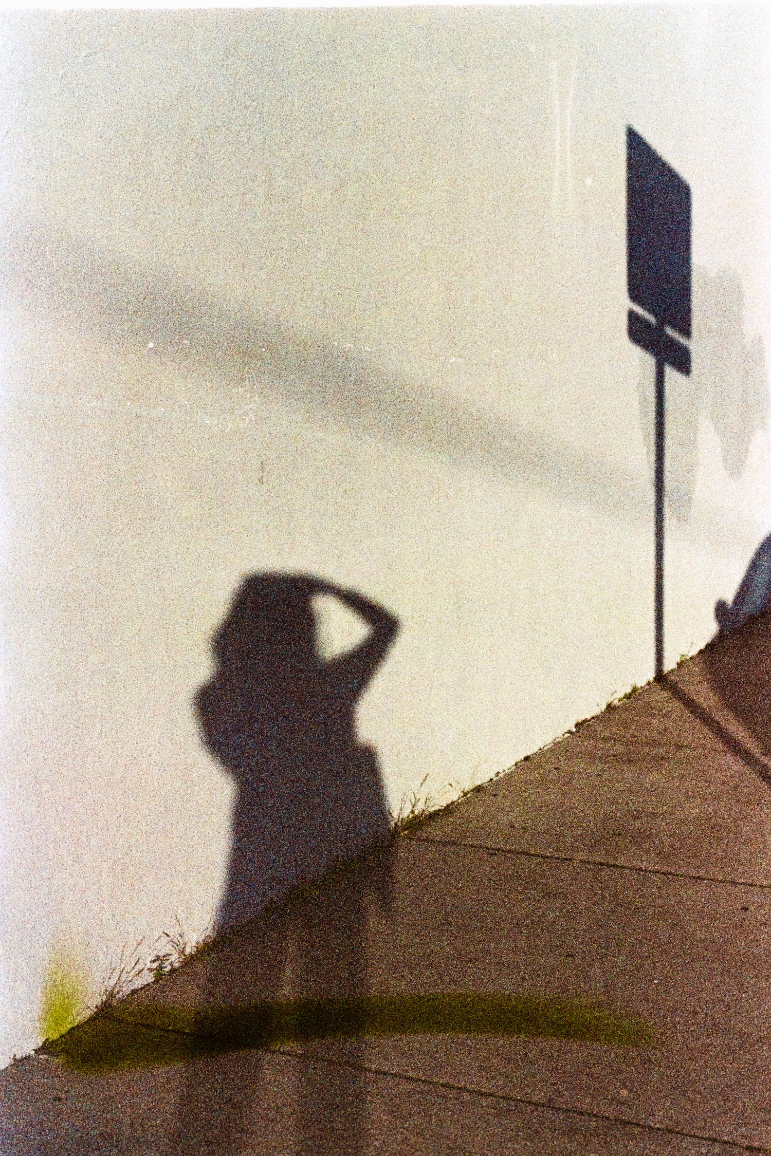 Shadow of a person shielding their eyes cast on a white wall beside a street sign post.