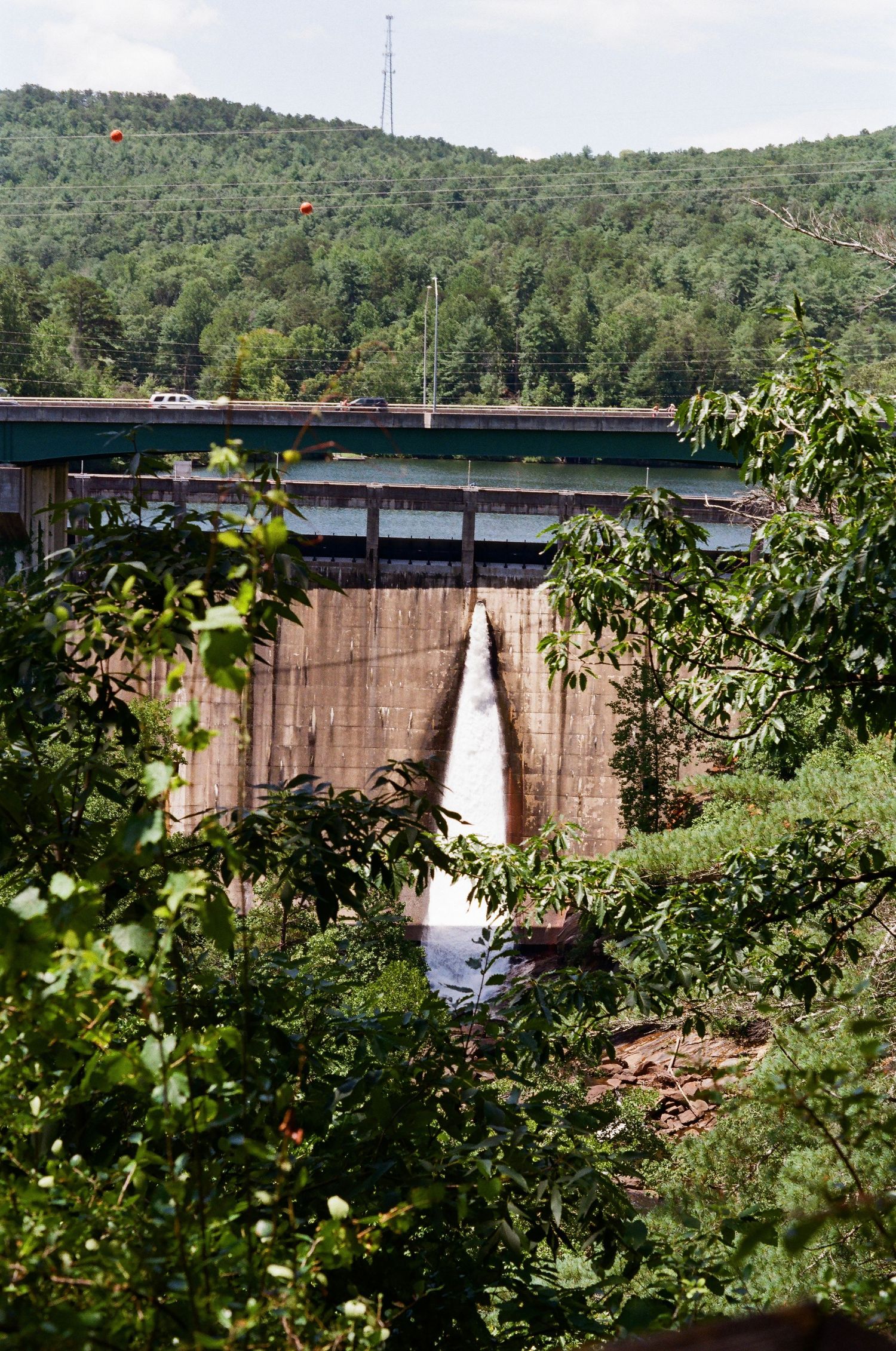 Water cascading down a concrete dam surrounded by lush green forest and a bridge on top under a cloudy sky.