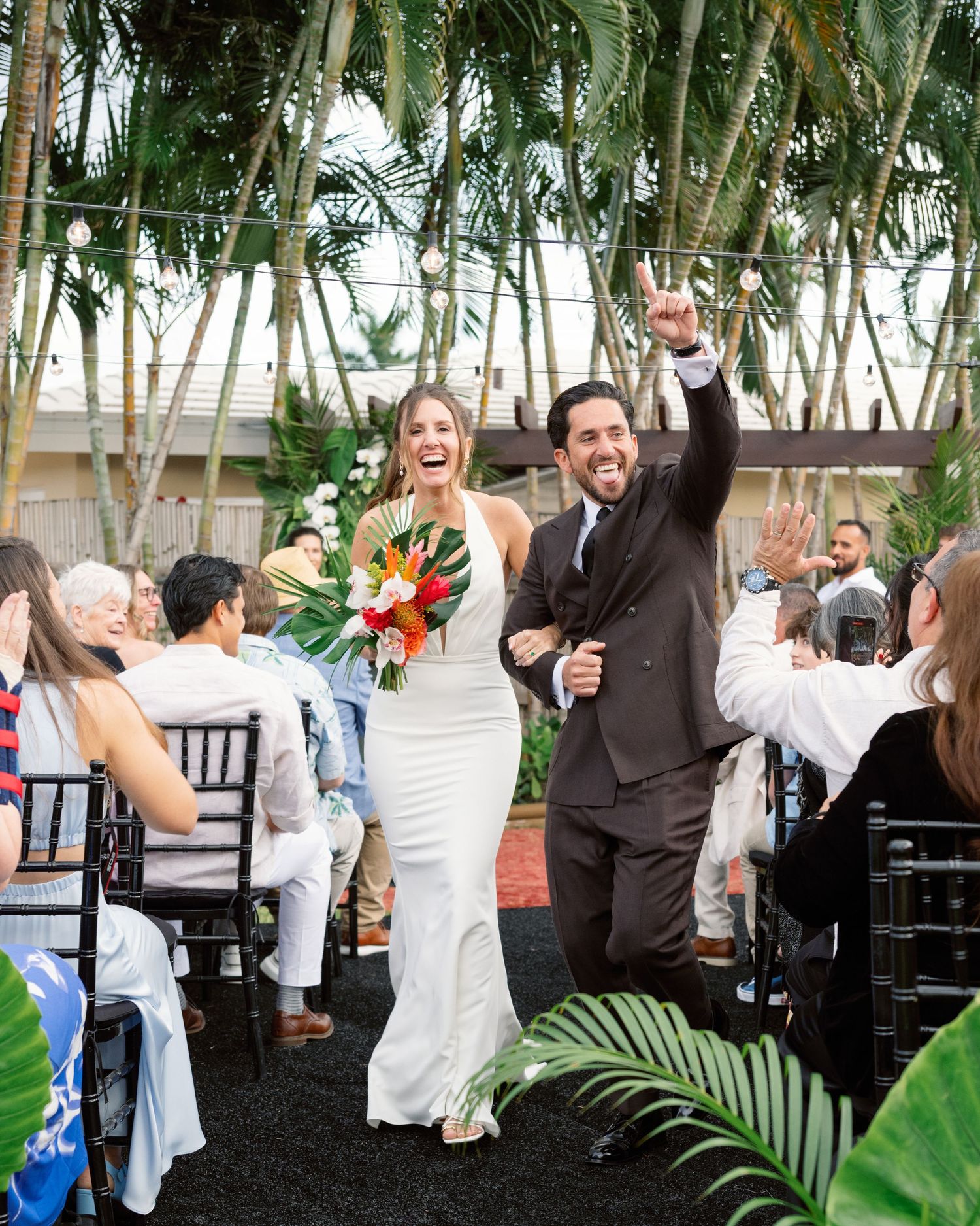 Newlyweds celebrate with guests during an outdoor tropical wedding ceremony with palm trees.