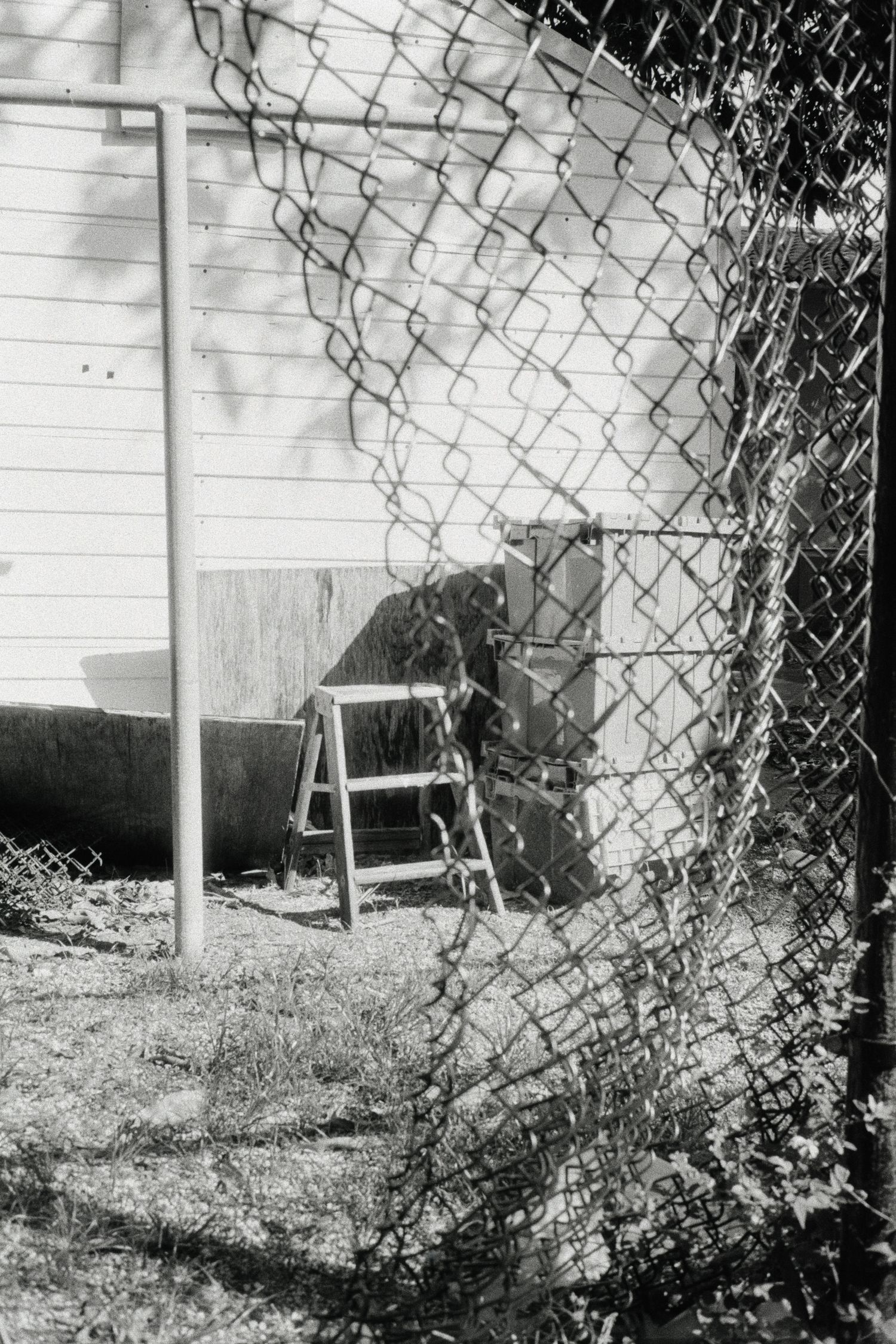 Black and white photo of a step ladder leaning against a white building behind a chain-link fence.