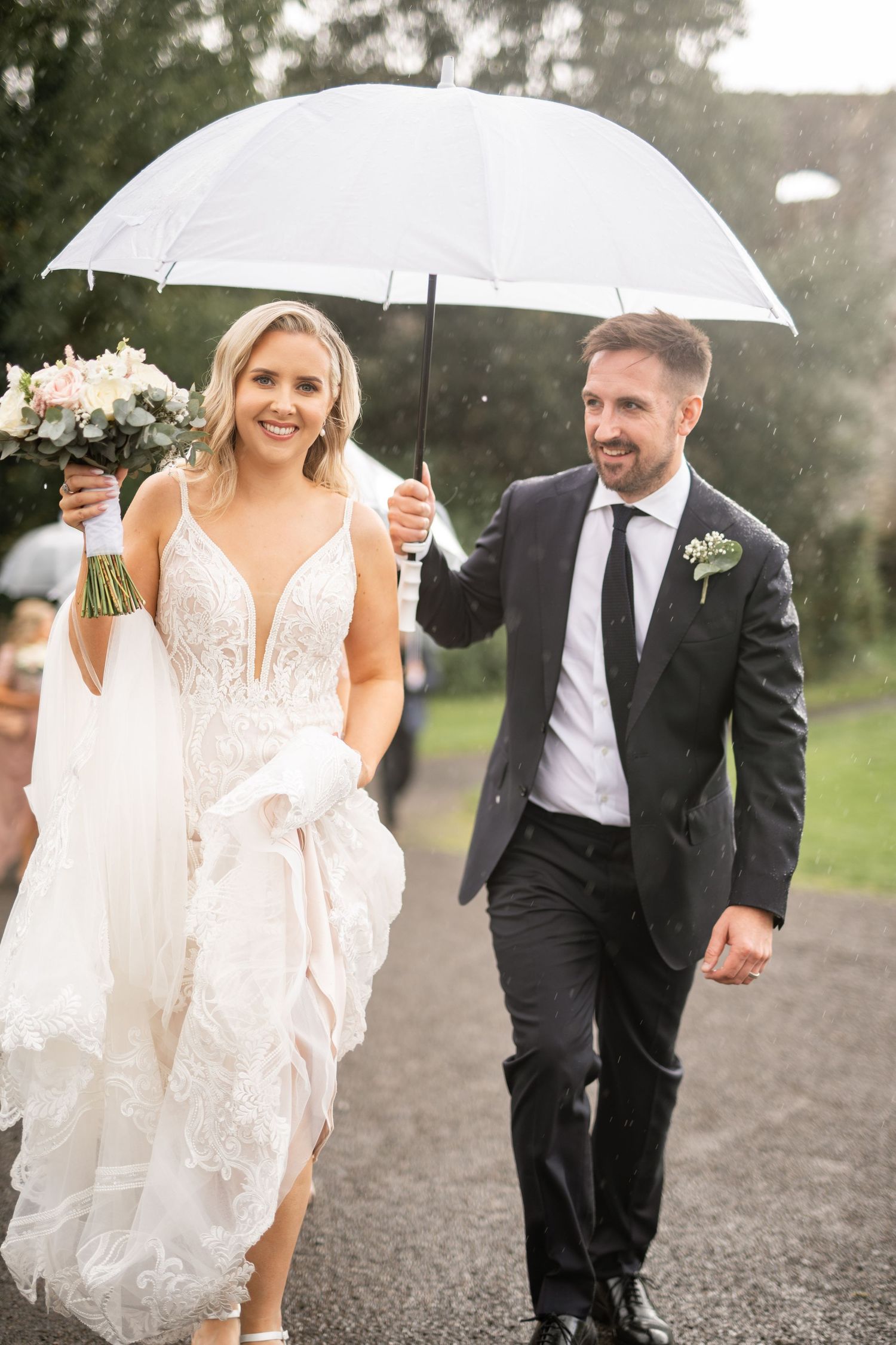 Happy bride and groom walk hand in hand under a white umbrella on a rainy wedding day.