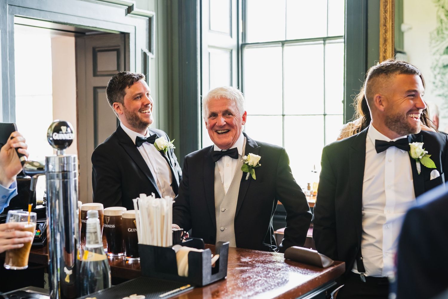Three men in black tuxedos smile and laugh together while standing at a wooden bar at a wedding.