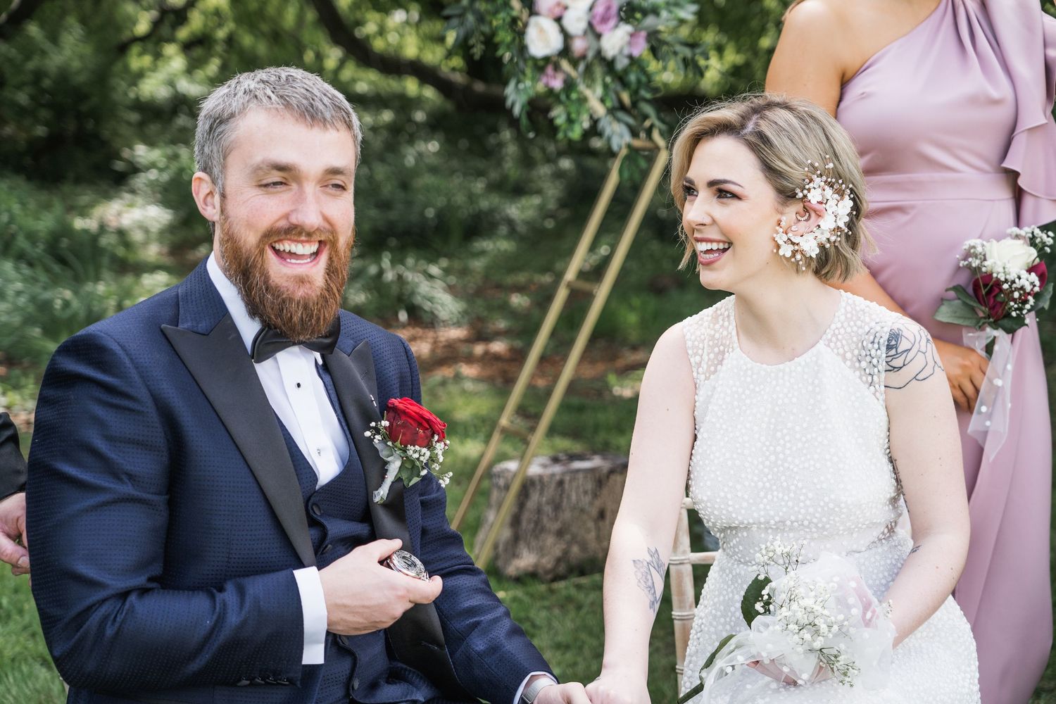Happy couple laughing during outdoor wedding ceremony, groom in navy suit with boutonniere, bride in elegant white gown.