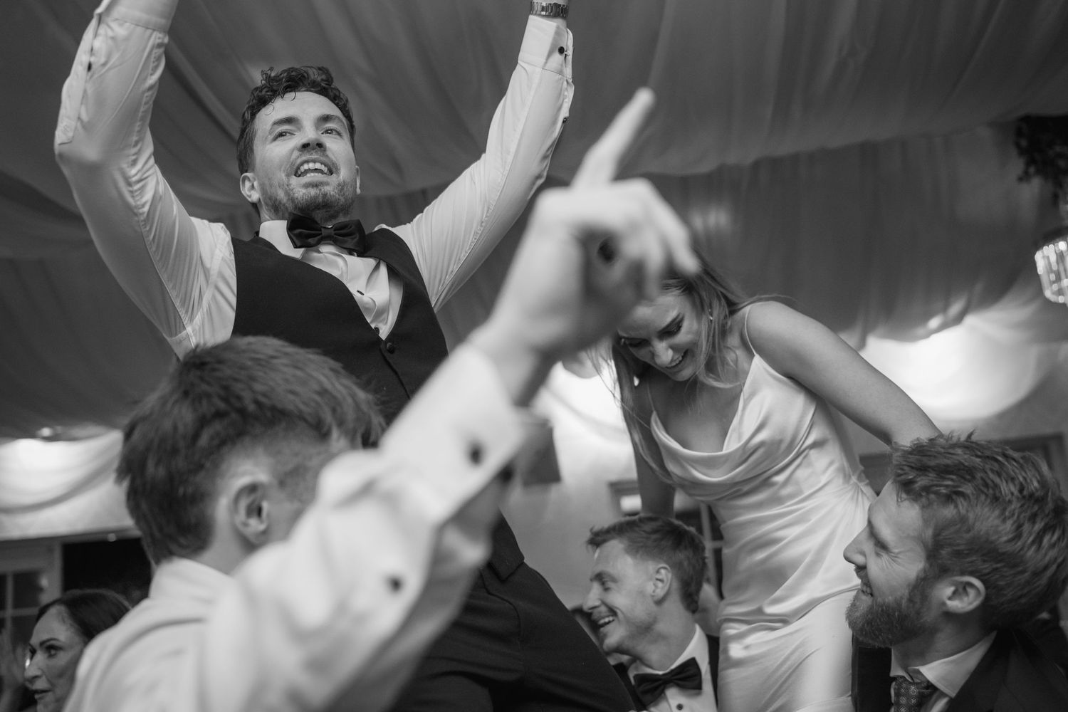 Groom raises arms in celebration while lifted by guests at festive wedding reception.