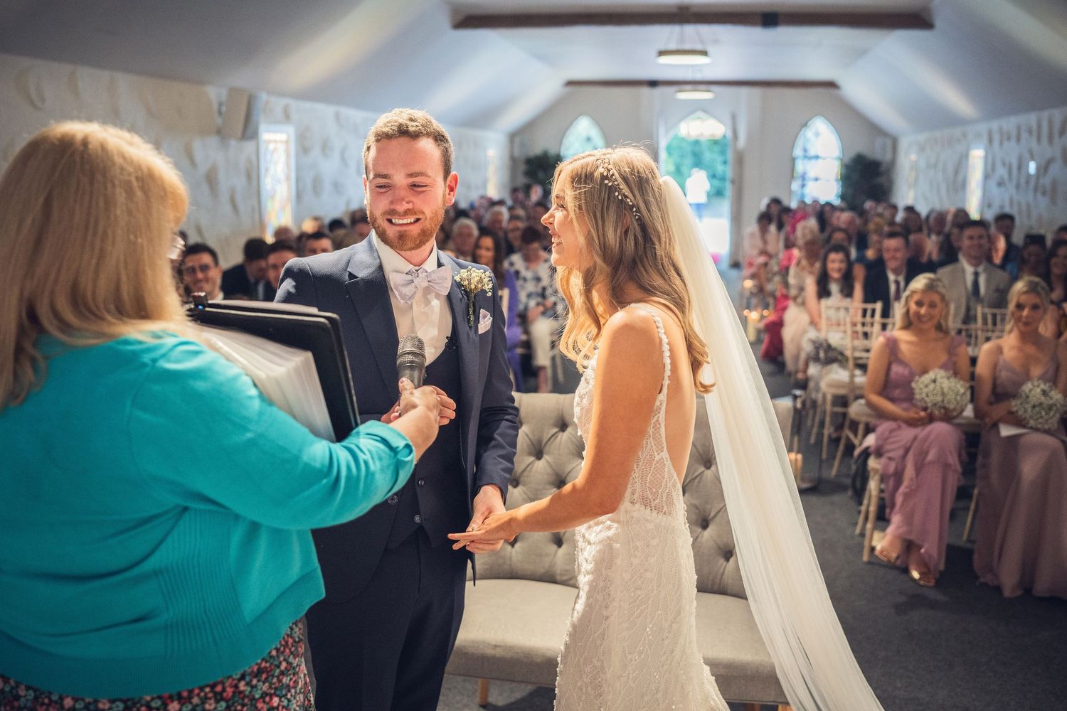 Groom smiles as officiant leads ring exchange ceremony in bright white chapel.