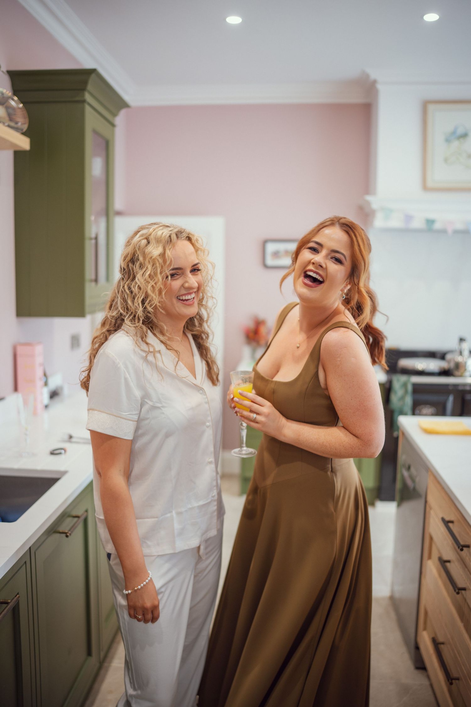 Two women laugh together in kitchen, one in white pajamas and one in olive bridesmaid dress holding champagne.