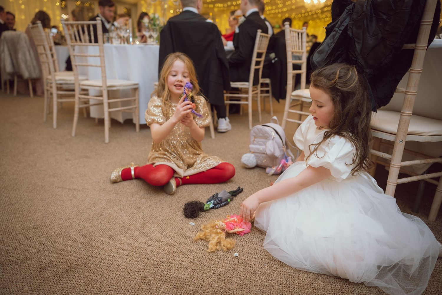 Two young flower girls playing with toys on the floor at a wedding reception.