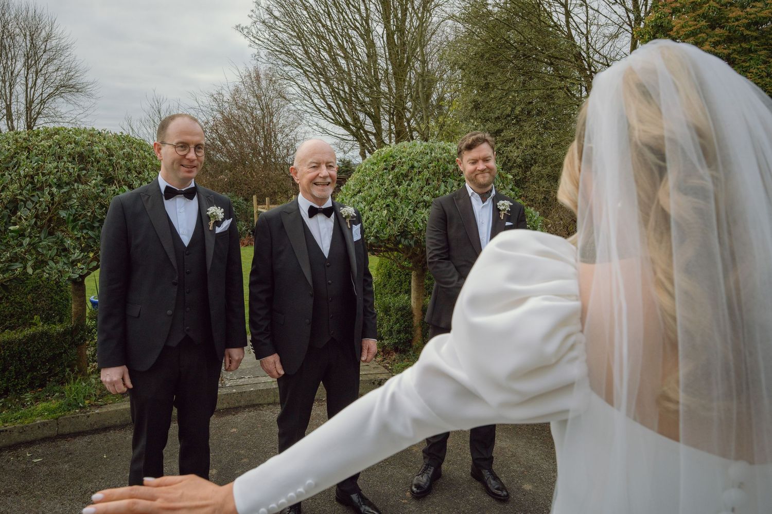 Bride in white gown and veil reaches toward three smiling groomsmen in tuxedos outdoors.
