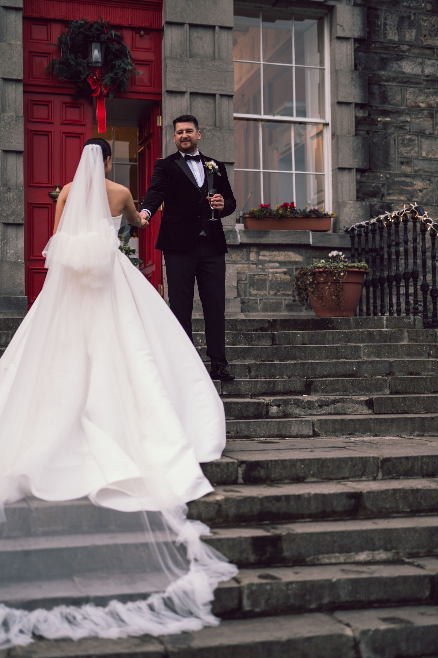 Bride in flowing white gown and veil walks up stone steps toward smiling groom in tuxedo at red door venue.