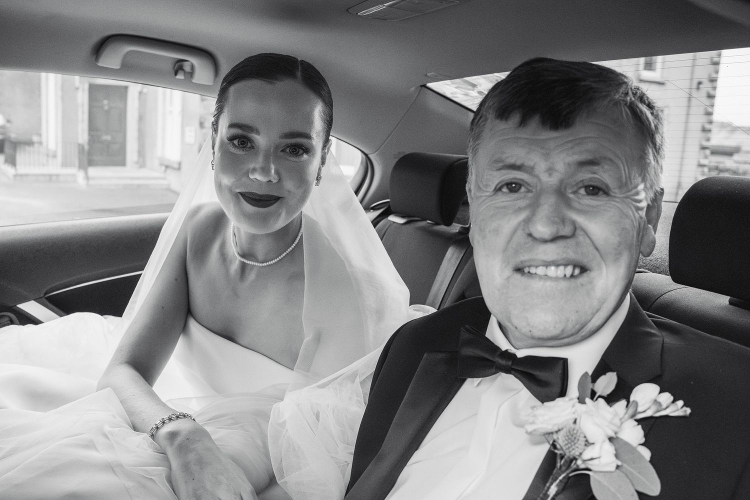 Bride and older gentleman smile together in car, black and white wedding photo before ceremony.