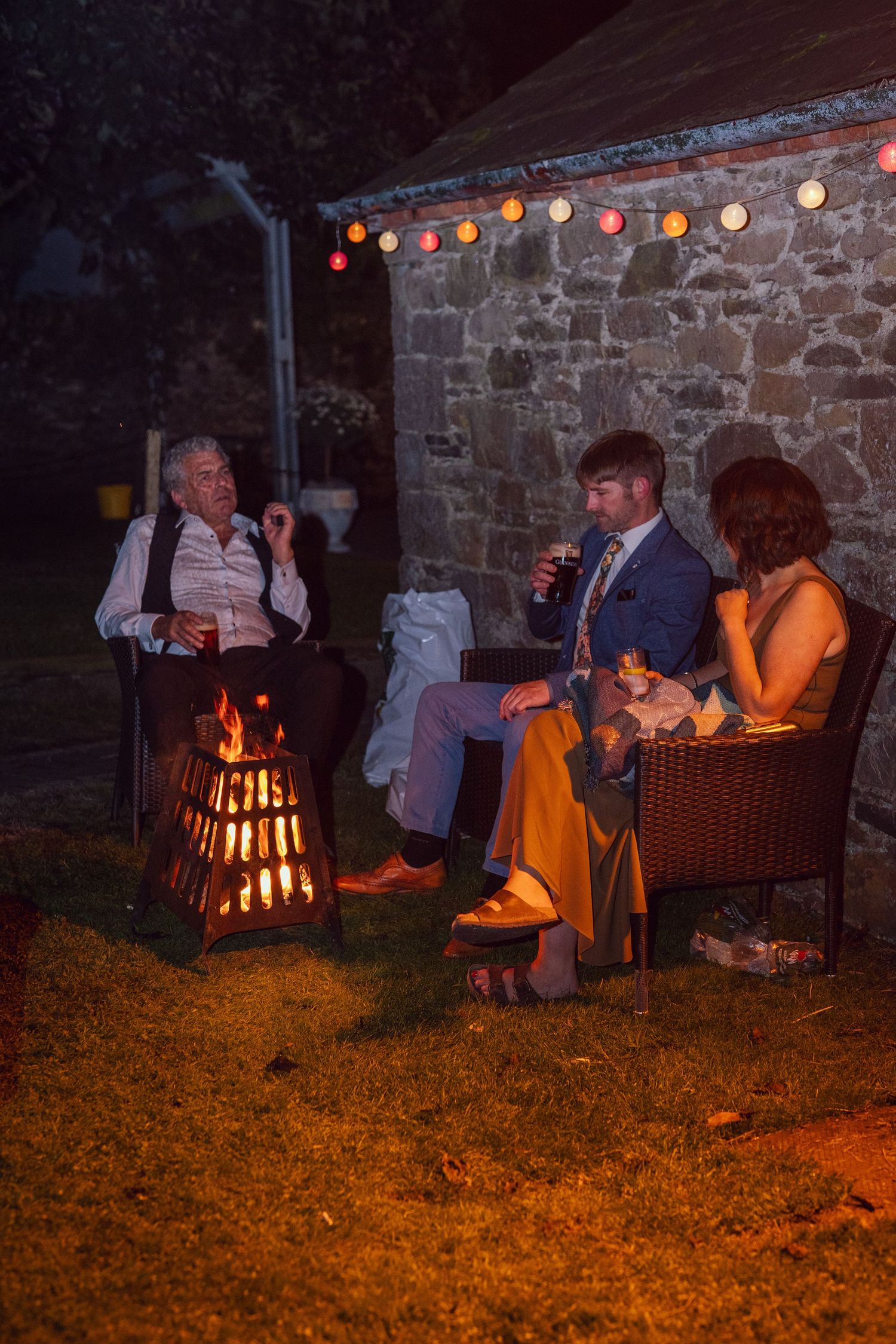 Three wedding guests relaxing around a warm outdoor fire pit at outdoor garden wedding reception in Co Meath.