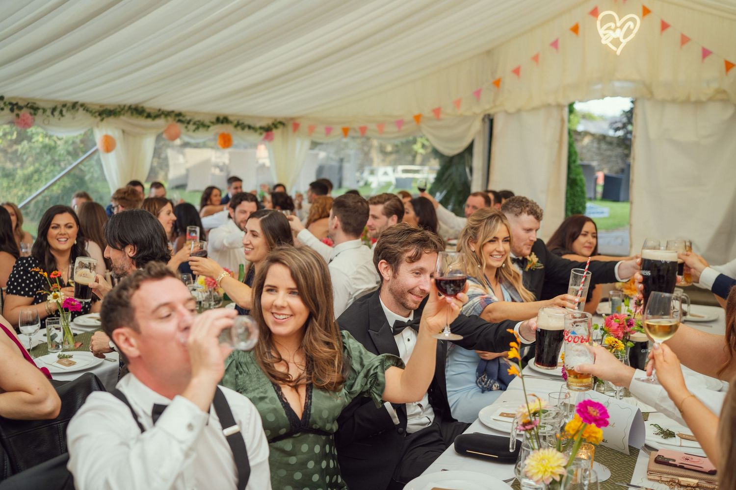 Wedding guests laughing and drinking wine at decorated tables inside a festive marquee reception at outdoor garden wedding reception in Co M