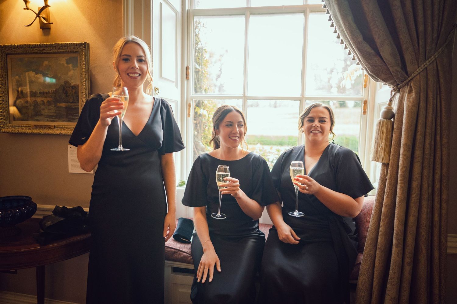 Three bridesmaids in matching black dresses raising champagne glasses in an elegant room at the Manor House Mount Juliet wedding Kilkenny.