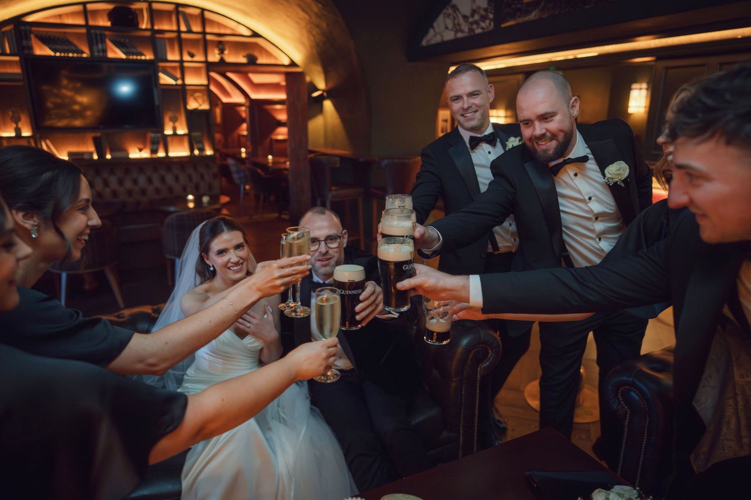 Wedding party toasting with champagne and beer glasses in a dimly lit elegant bar venue at the Manor House Mount Juliet wedding Kilkenny.
