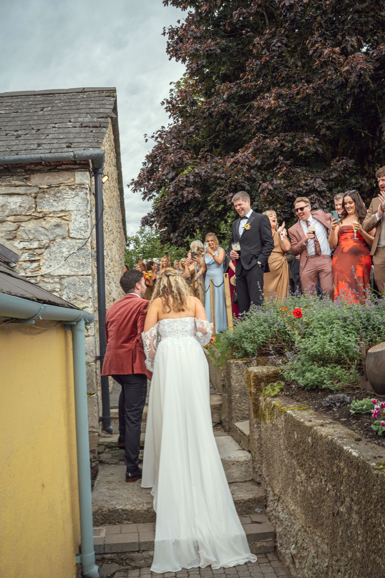Bride in white gown walks toward groom and wedding guests applauding on stone steps at outdoor venue.