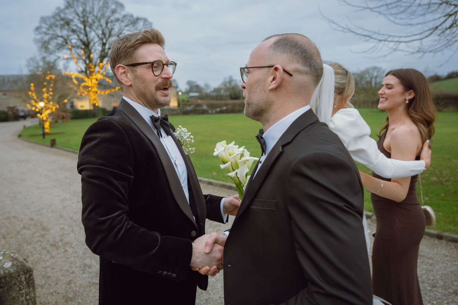 Two men in tuxedos shake hands warmly outside a venue as bride and bridesmaid chat in background.