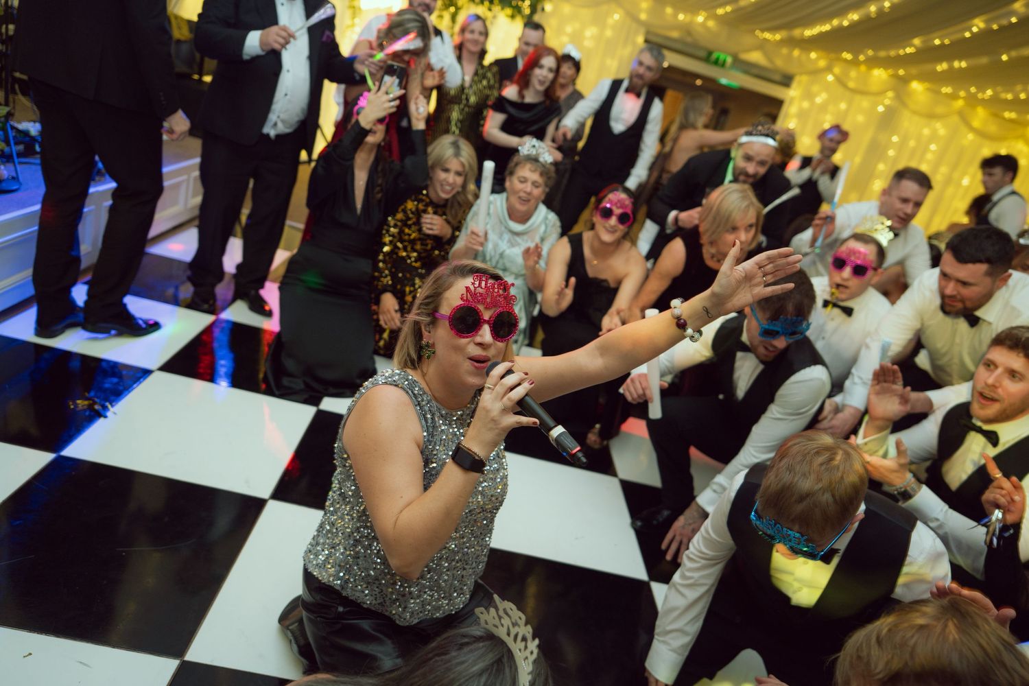 A woman in a sequin dress sings on a checkered dance floor surrounded by cheering wedding guests.