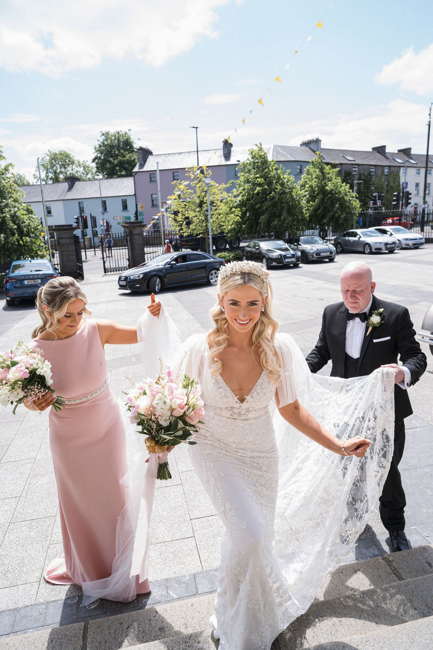 Bride in lace wedding gown walks up cathedral steps with bouquet assisted by family members.
