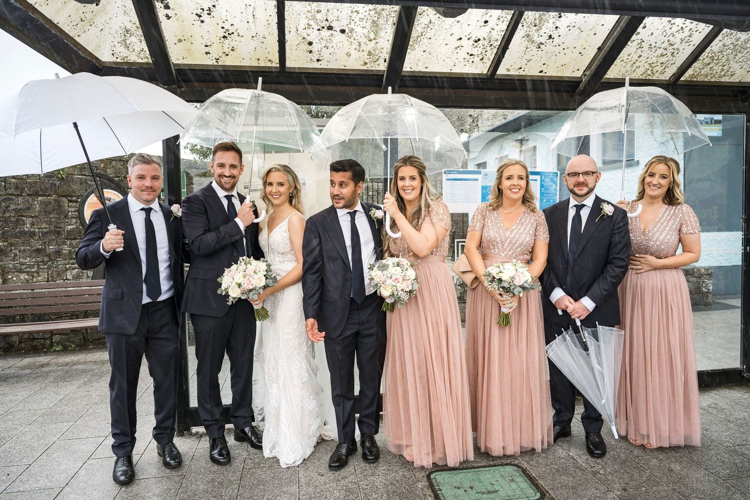 Wedding party poses with clear umbrellas in the rain, bridesmaids in dusty rose gowns with bouquets.