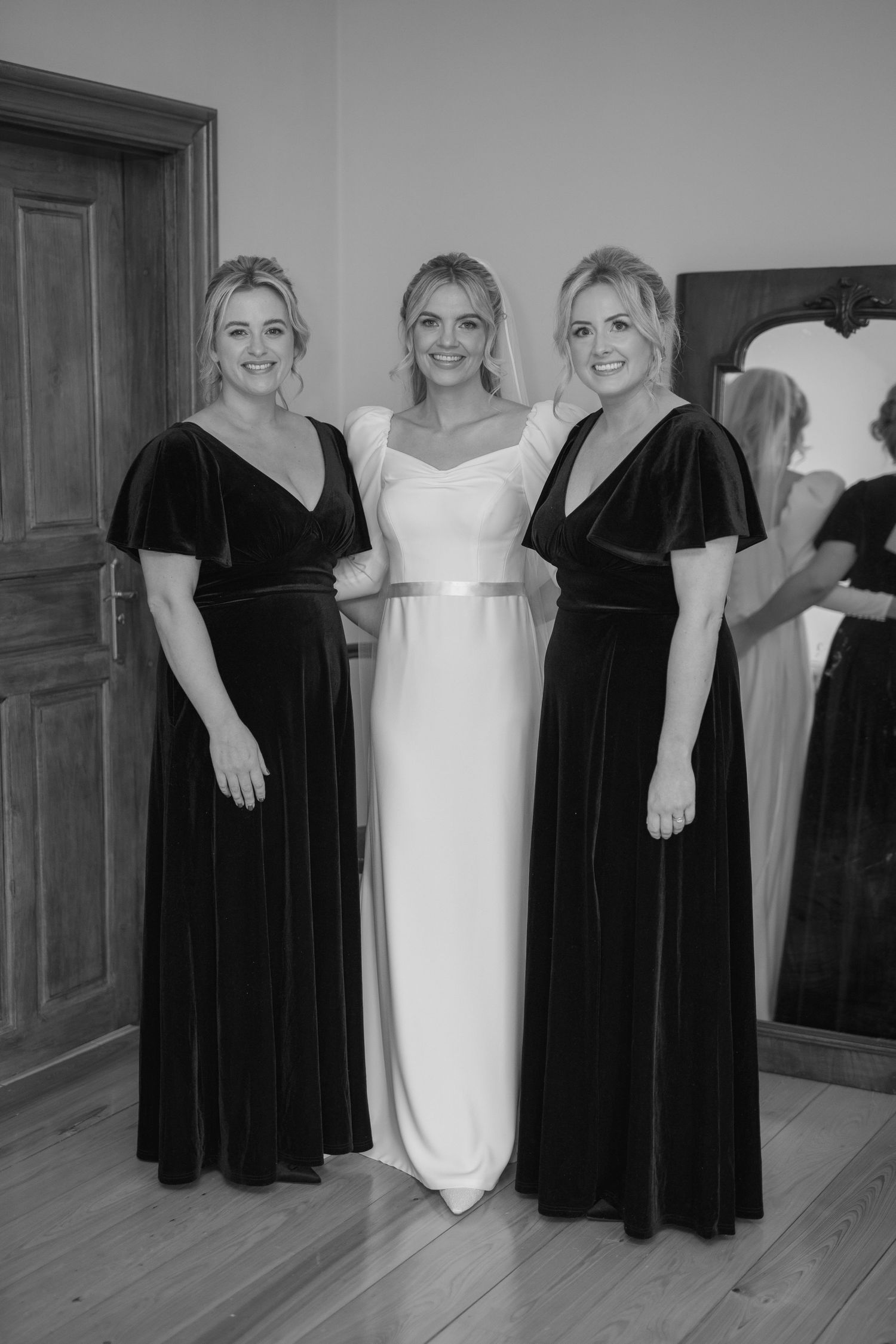 Bride in white gown poses with two bridesmaids in dark velvet dresses before wedding.