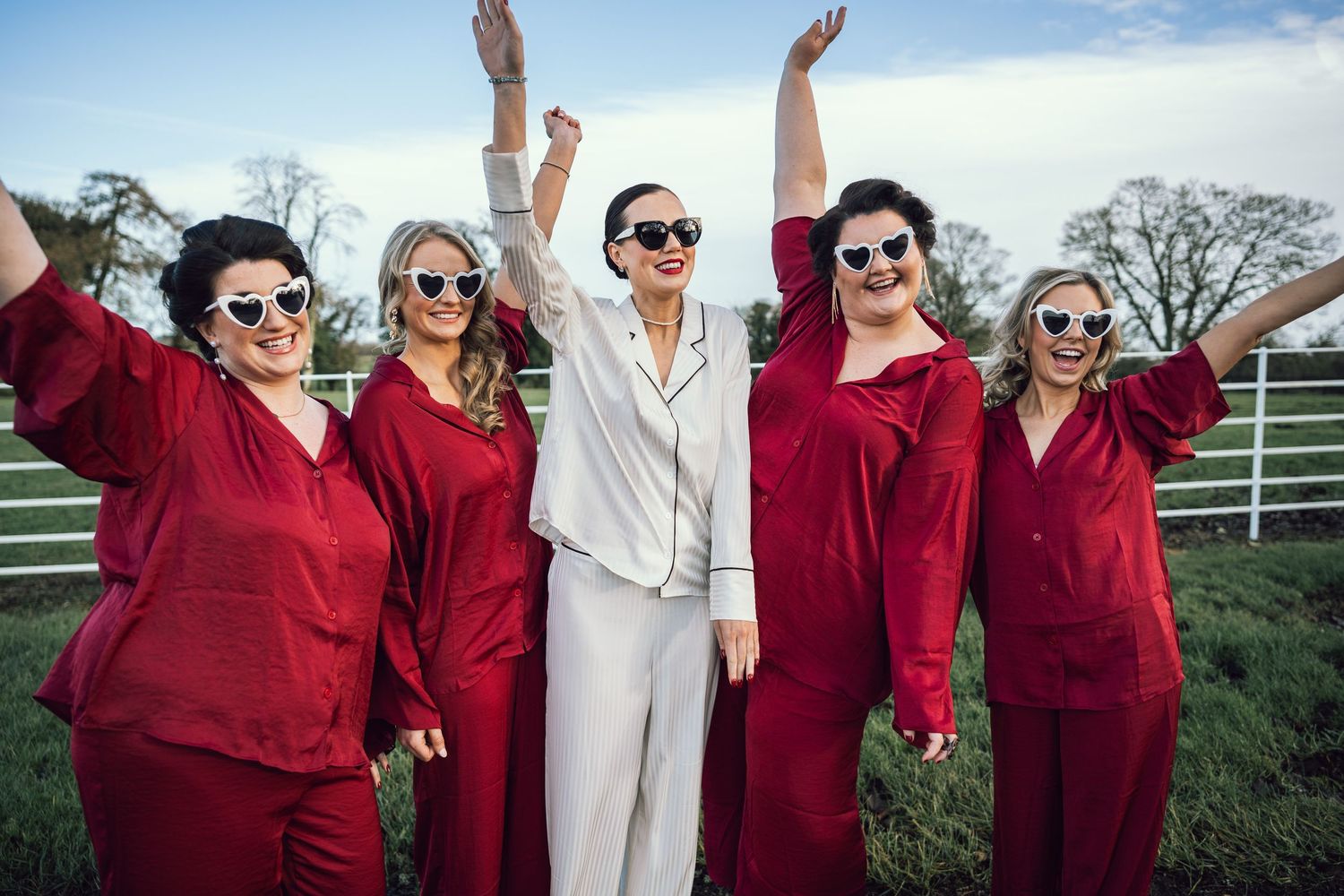 Bride in white pajamas celebrating with four bridesmaids in matching red pajamas outdoors.