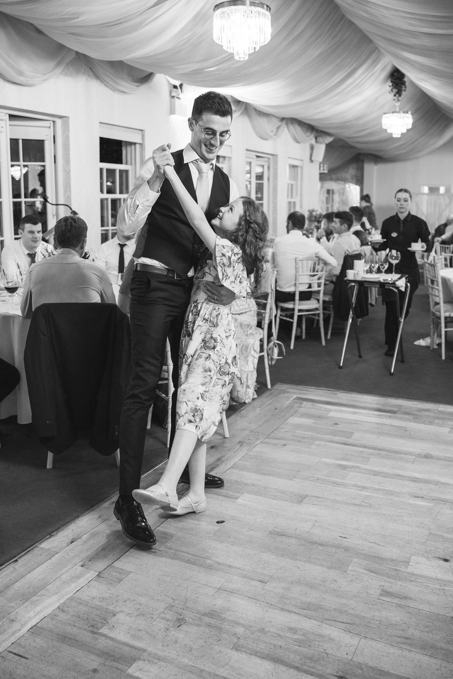 Black and white photo of groom dancing with young girl on wooden dance floor at wedding reception.