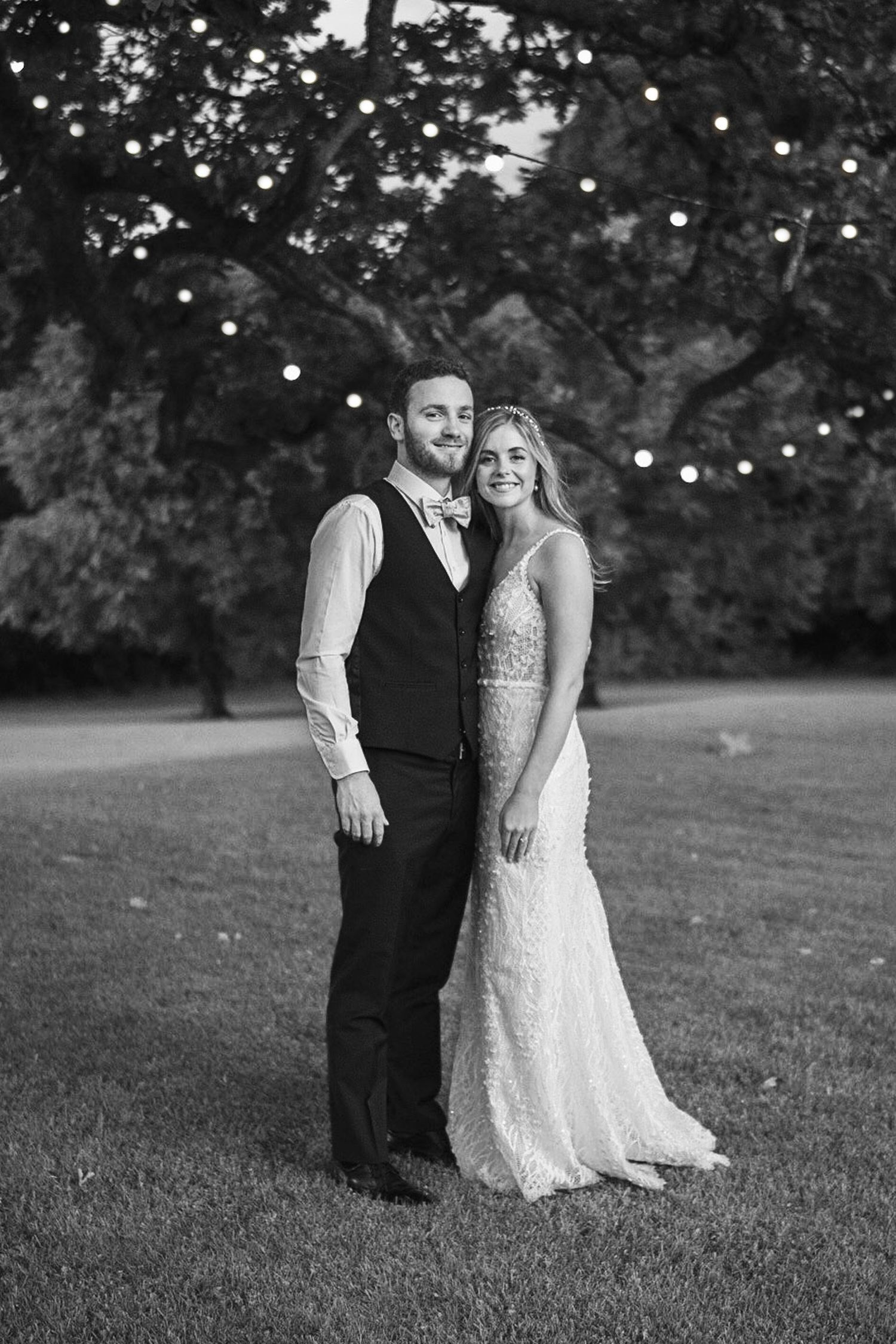 Black and white portrait of a smiling bride and groom standing together outdoors with fairy lights in trees behind.