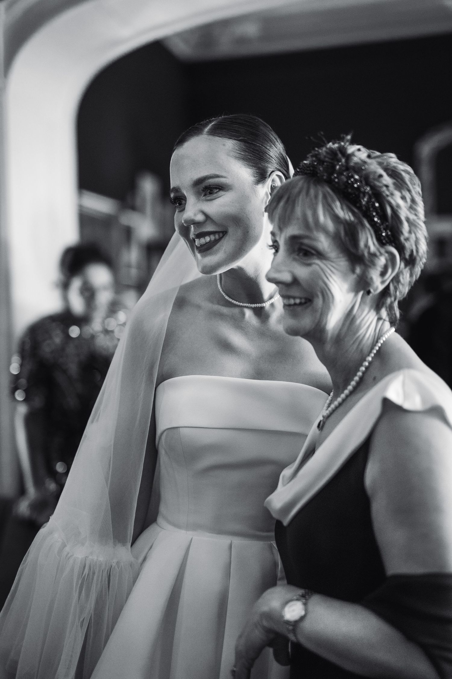 Black and white photo of smiling bride in strapless gown and veil posing with older woman in pearl necklace.