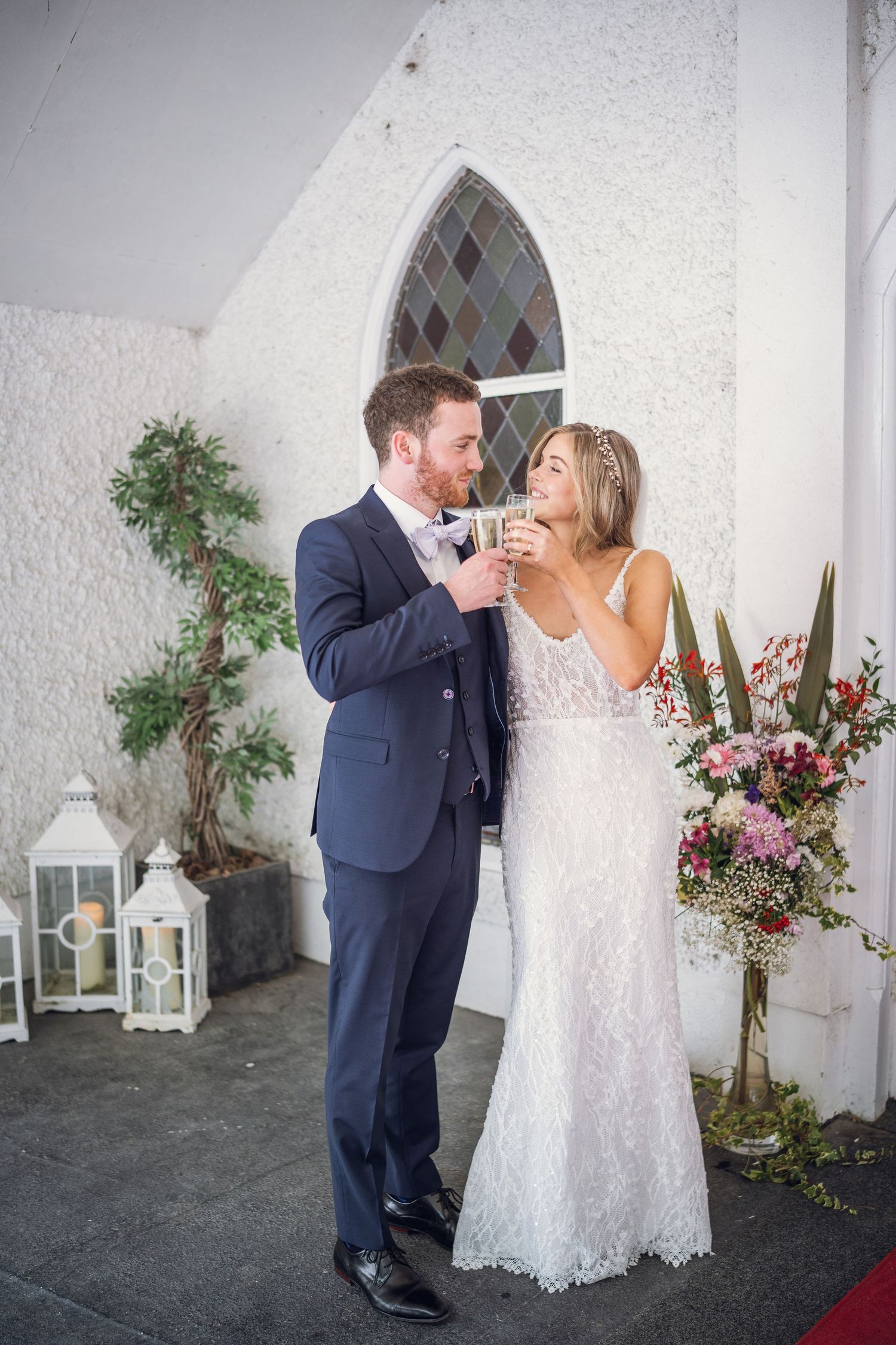 Newlyweds share champagne toast outside white church decorated with lanterns and pink floral arrangements.