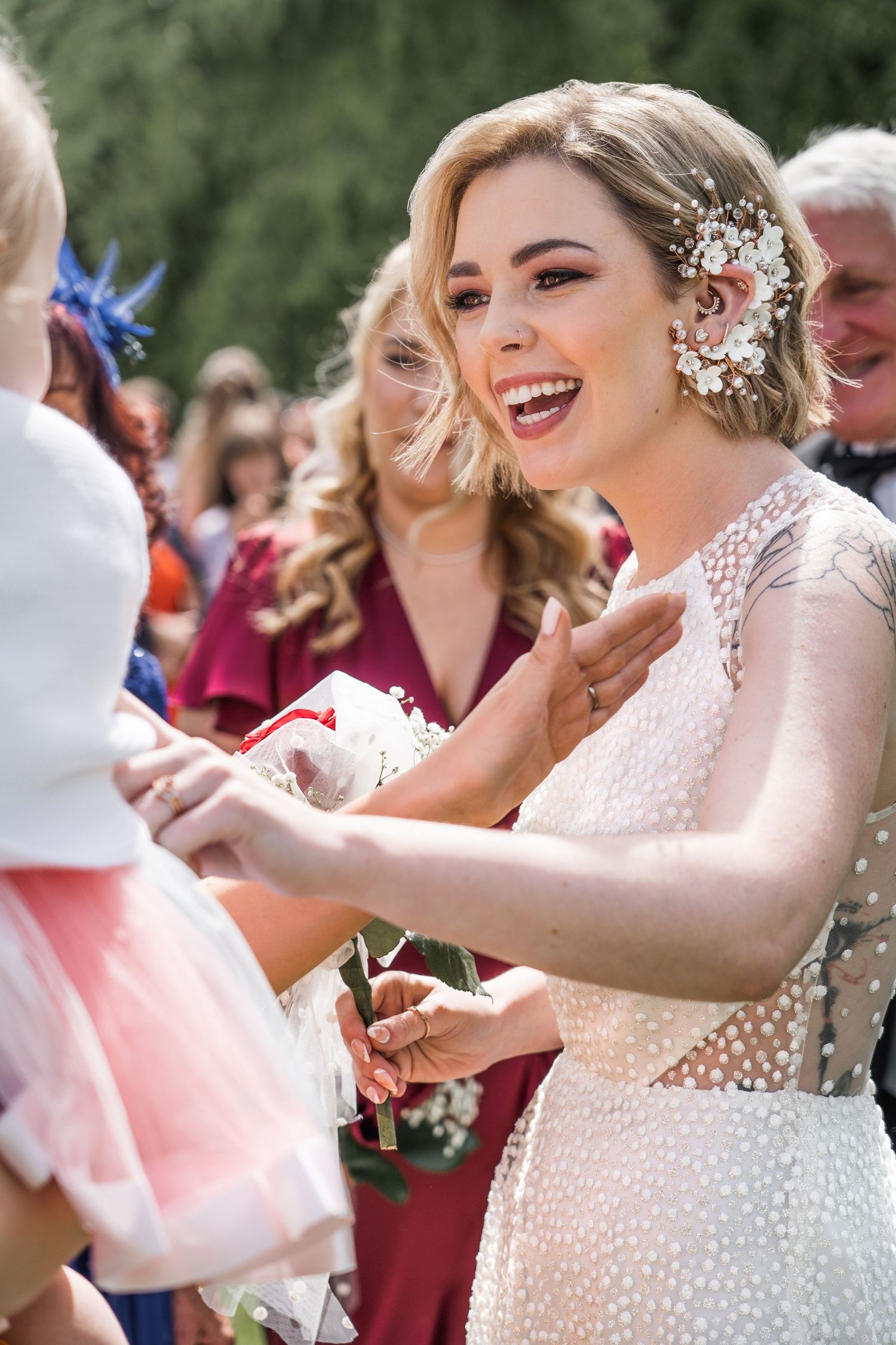 Joyful bride in pearl-embellished gown with floral hair accessory laughing while greeting guests at outdoor wedding.