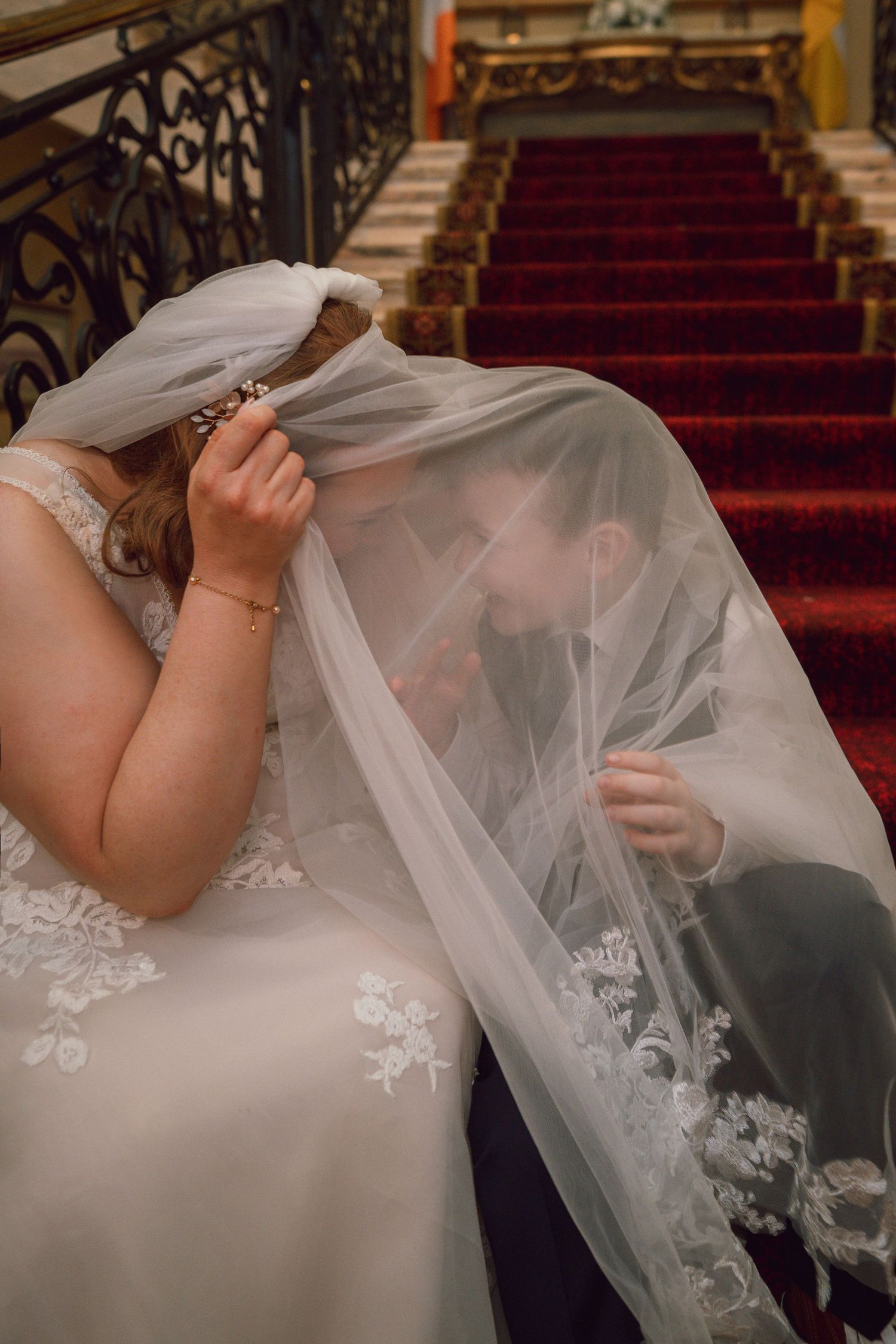 Bride and groom sharing intimate moment hidden under lace wedding veil on grand staircase.