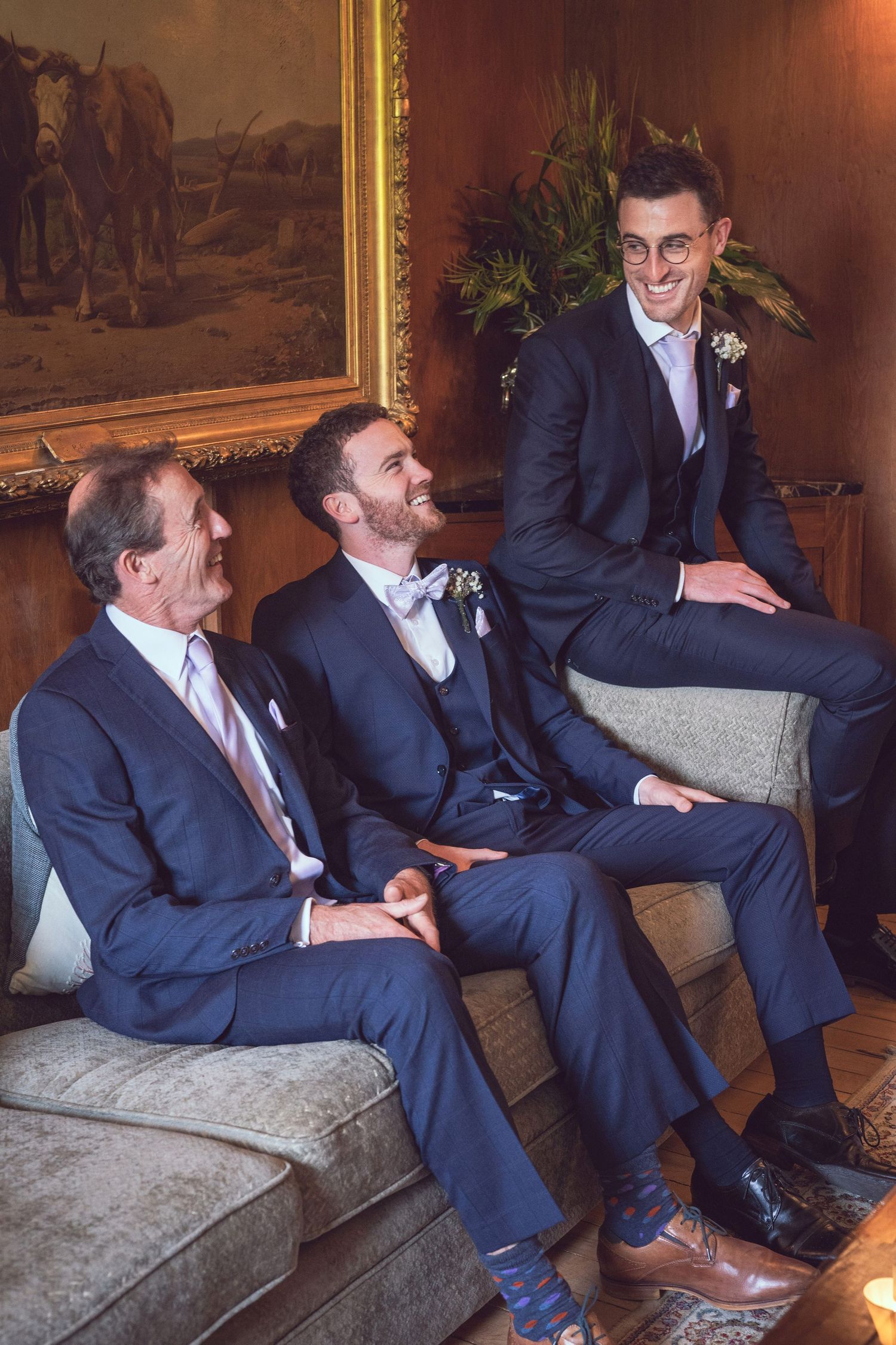 Three men in navy suits laugh and relax on a sofa before a wedding ceremony in an elegant room.