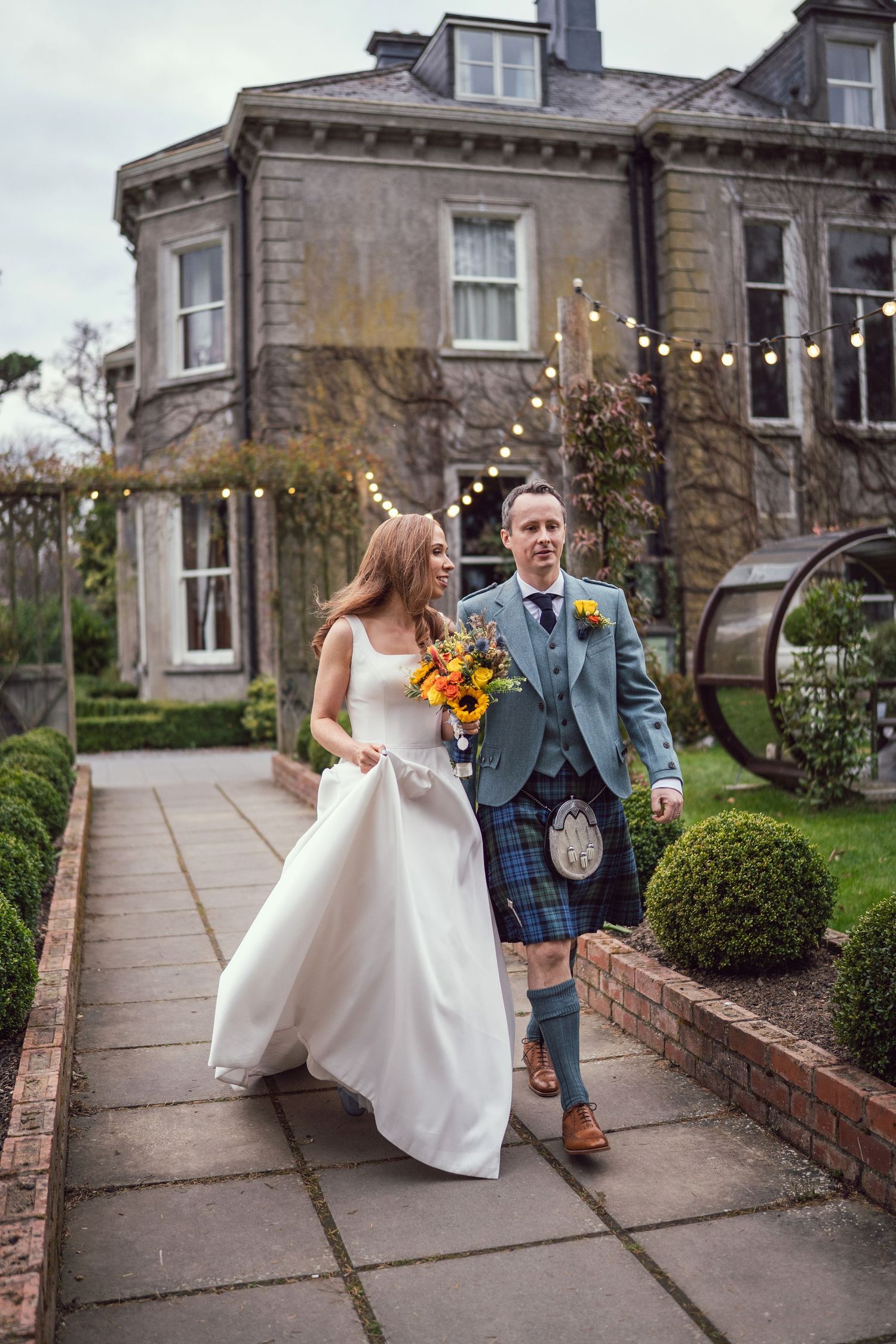 Bride and groom in Scottish kilt walking hand-in-hand outside a grand stone manor with fairy lights.