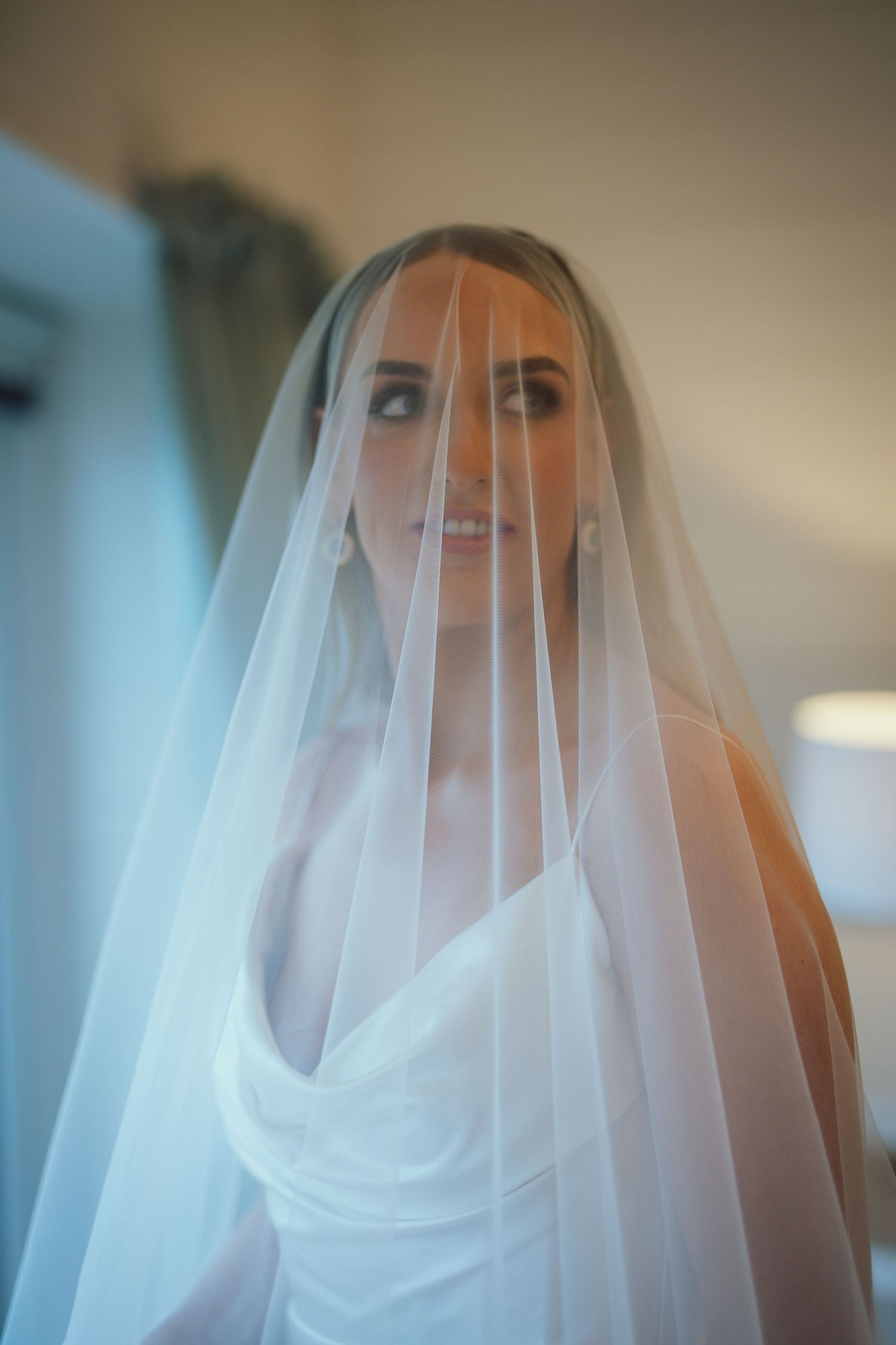 Bride wearing a white wedding dress and sheer veil draped over her face, posing elegantly in a softly lit room.