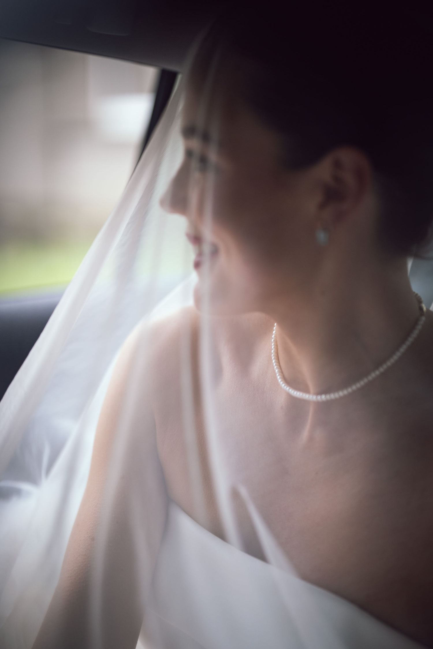 Bride in white wedding dress and pearl necklace gazing out car window, soft veil draped over her shoulder.