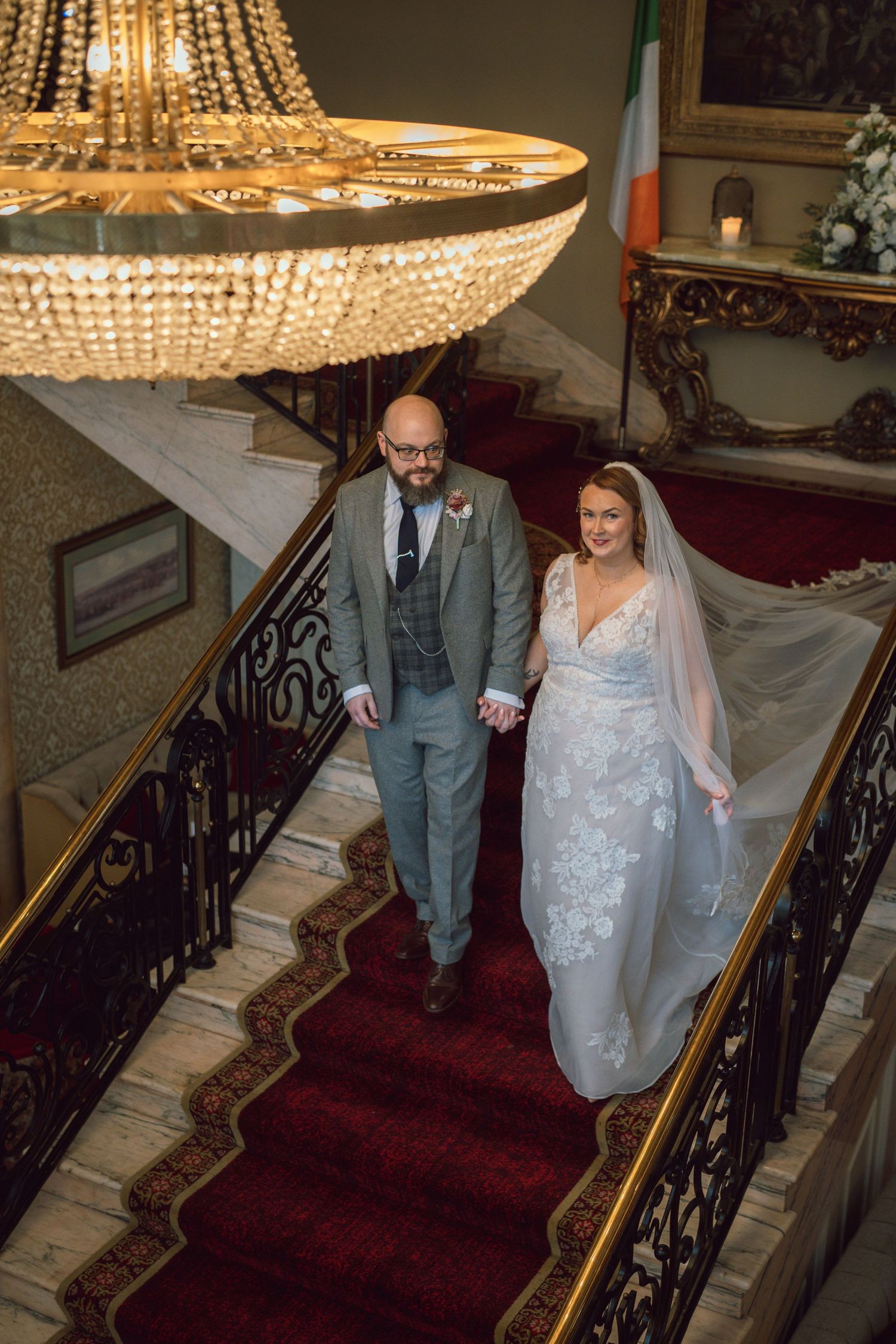 Bride and groom descending grand staircase beneath crystal chandelier with Irish flag in background.