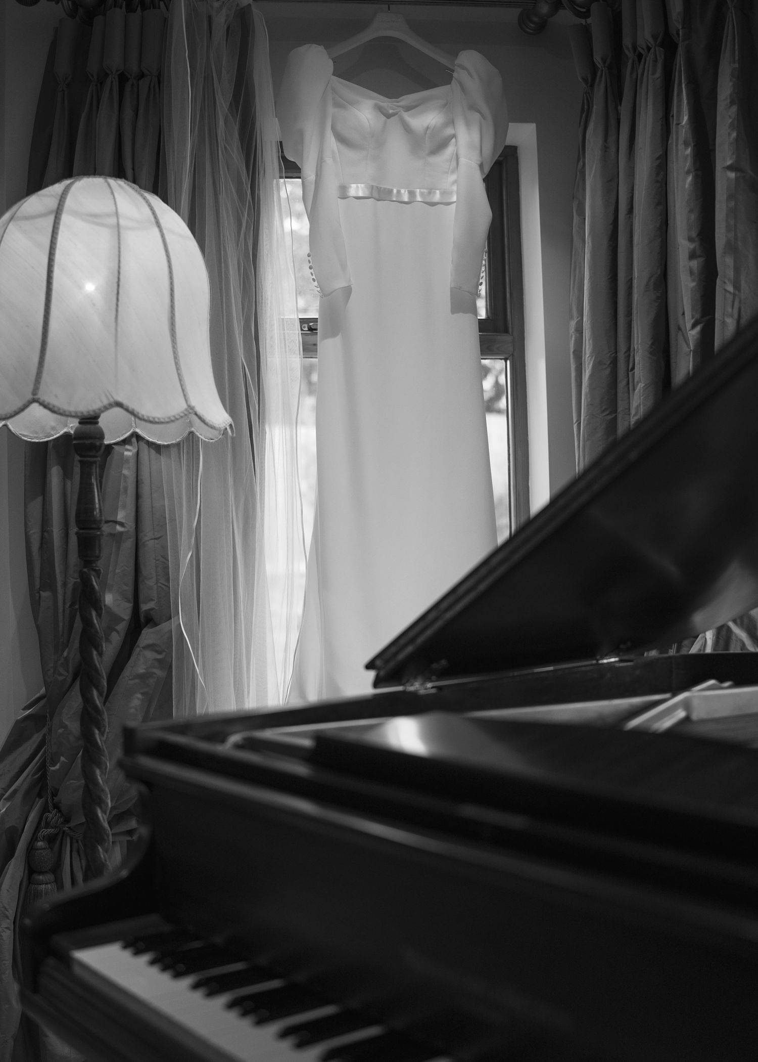 Elegant wedding dress hanging by window above grand piano in black and white bridal preparation photo.