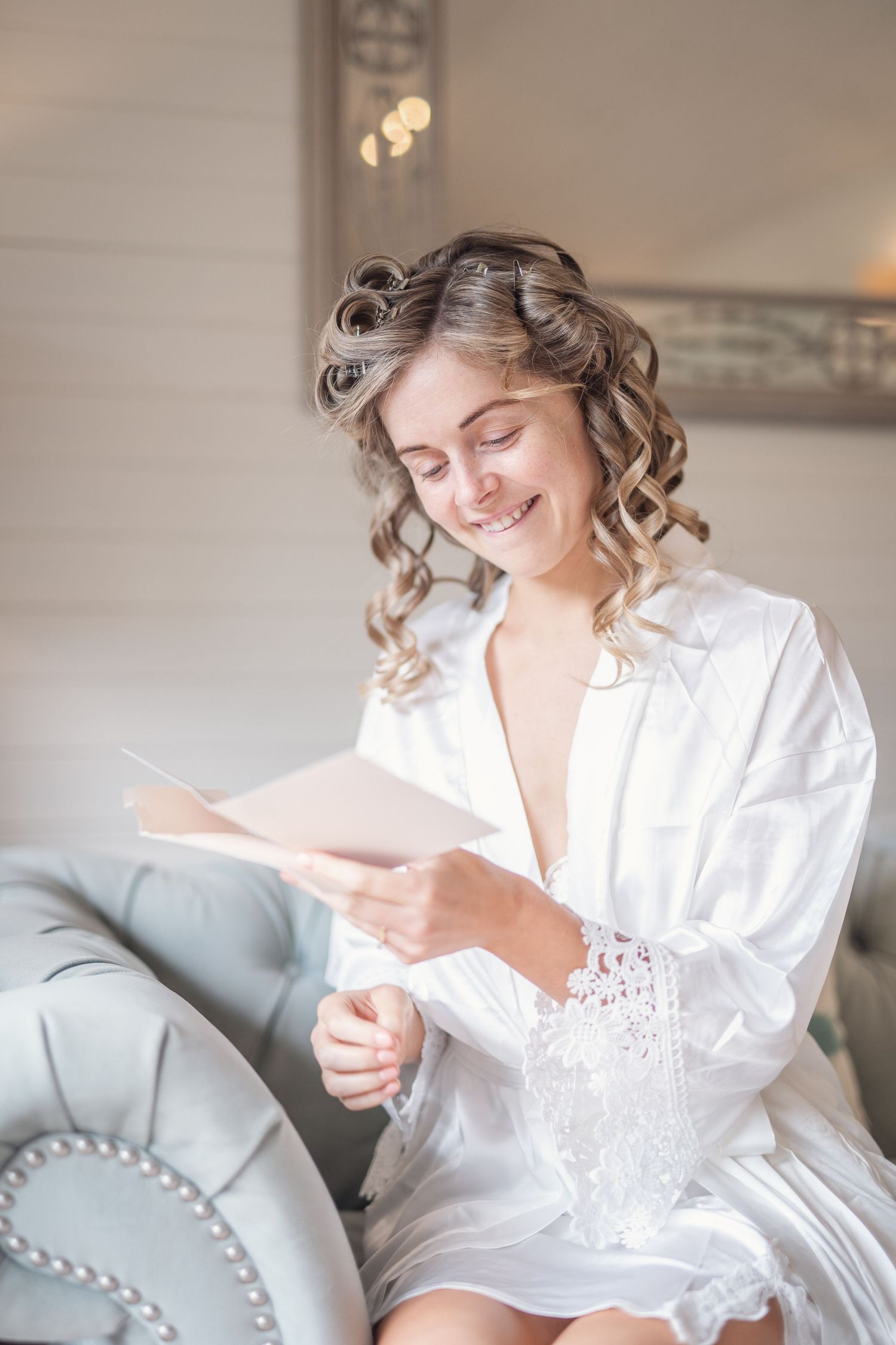 Smiling bride in white robe reading a card while getting ready on her wedding morning.