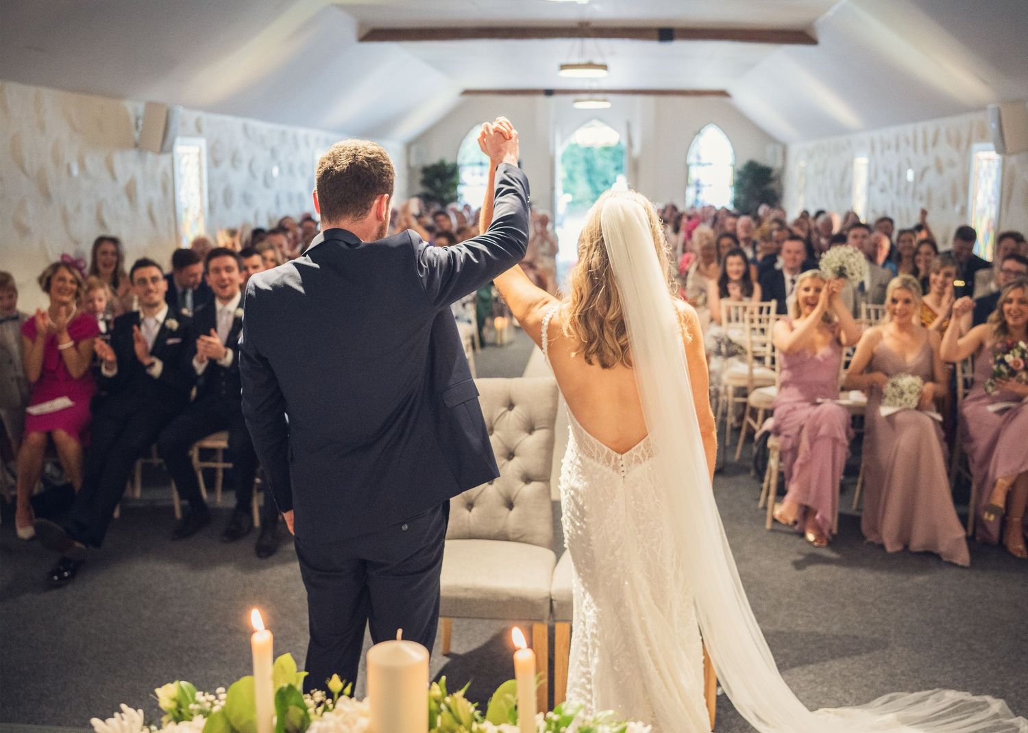 Newlyweds celebrate with raised fists before cheering guests in a bright white wedding chapel.