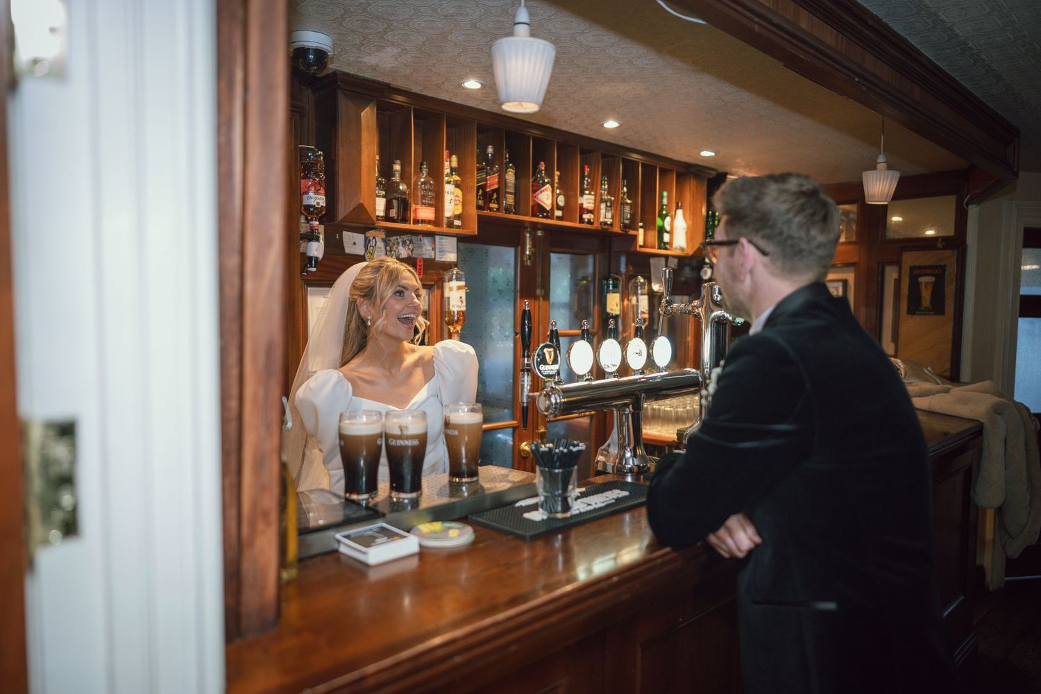 Bride in white gown serving Guinness pints to groom at a traditional wooden bar.