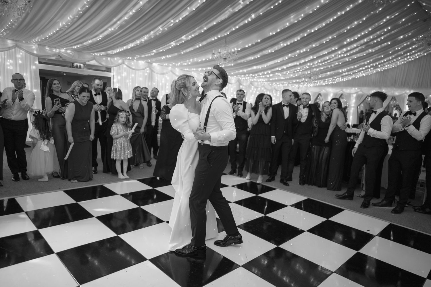 Bride and groom sharing first dance on checkered floor surrounded by wedding guests.